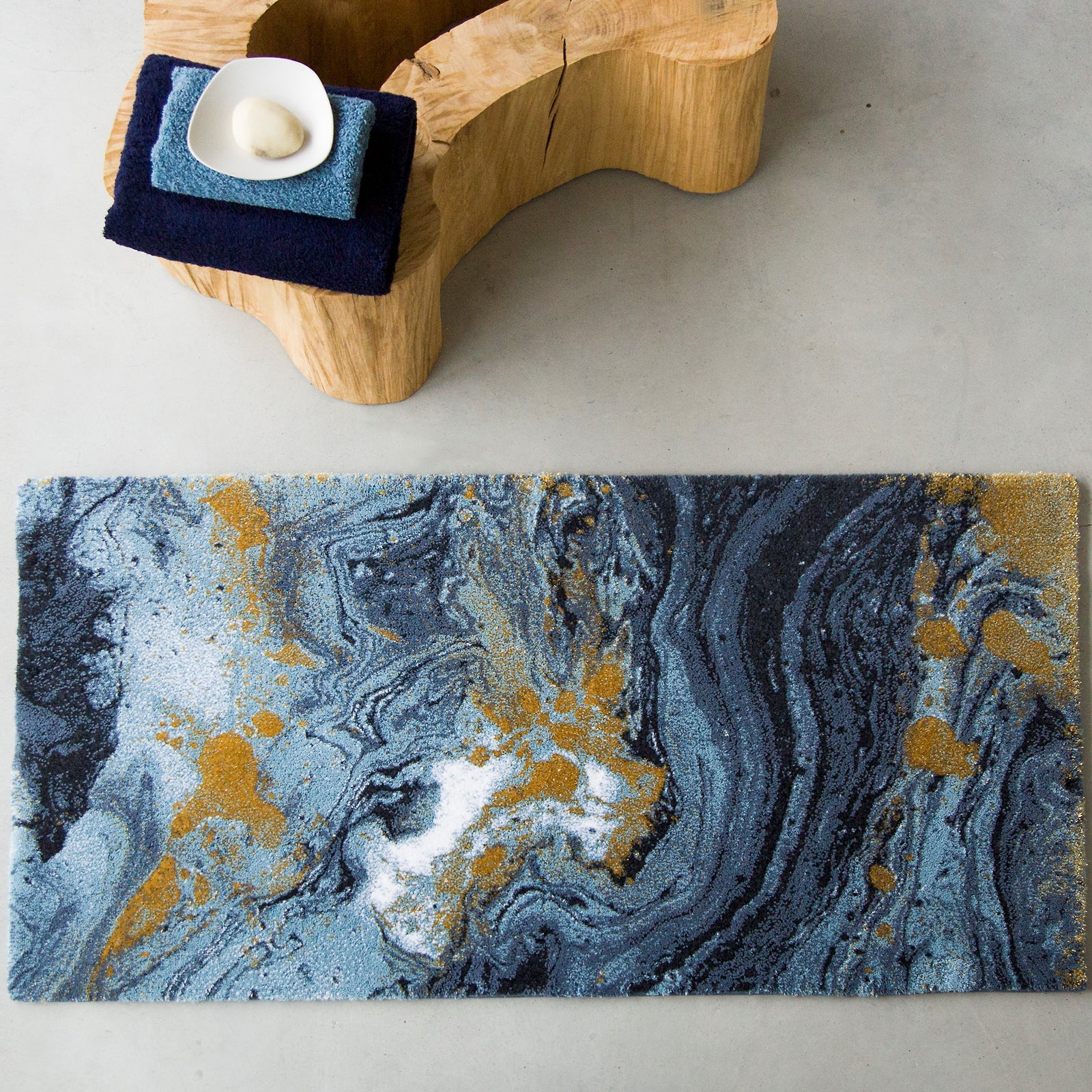 A rectangular rug with a marbled pattern in shades of blue, white, black, and gold is placed on a light gray floor next to a wooden stool with folded towels and a small dish on top.