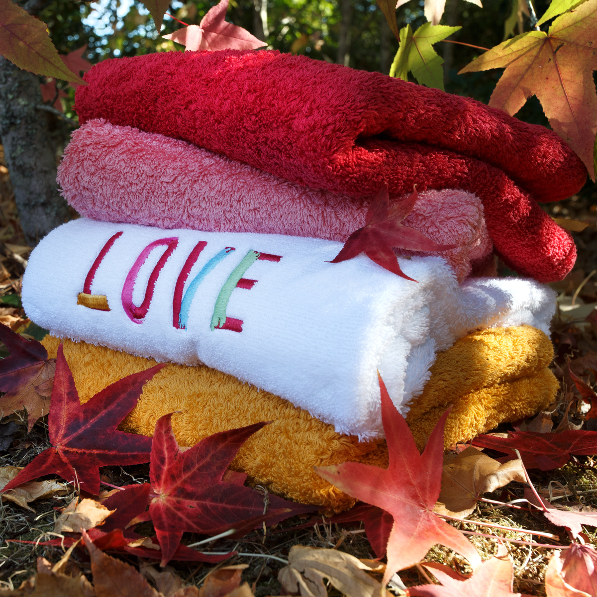 Four folded towels, including an embroidered LOVE towel, with autumn leaves scattered around them outdoors.