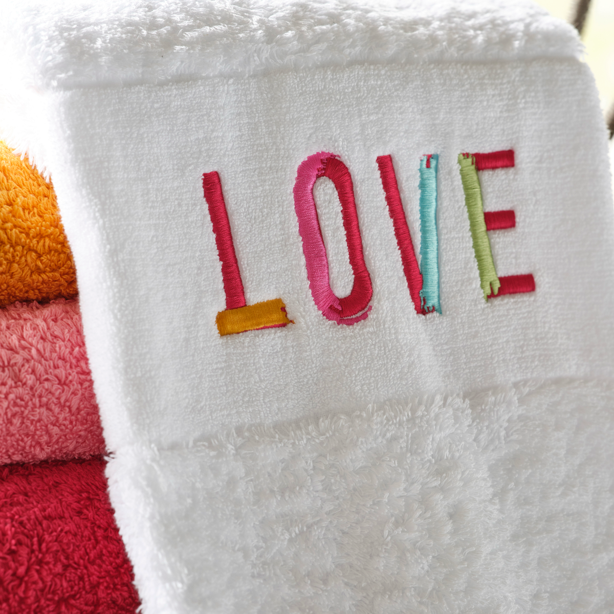An embroidered LOVE towel made of Egyptian cotton is draped over a stack of colorful towels.