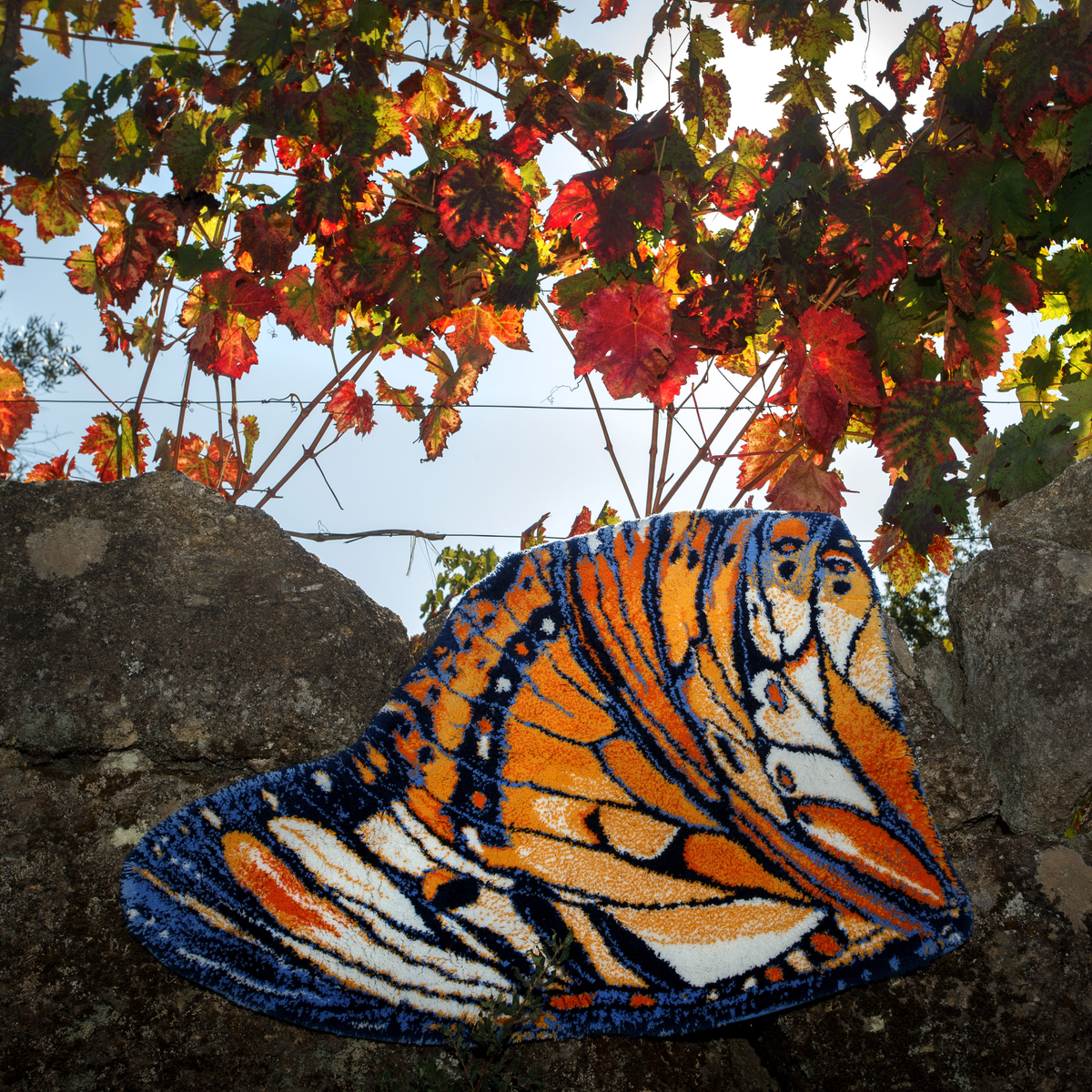 An Abyss Habidecor Mariposa Bath Rug, shaped like a butterfly wing, drapes over a stone wall under sunlit grapevines