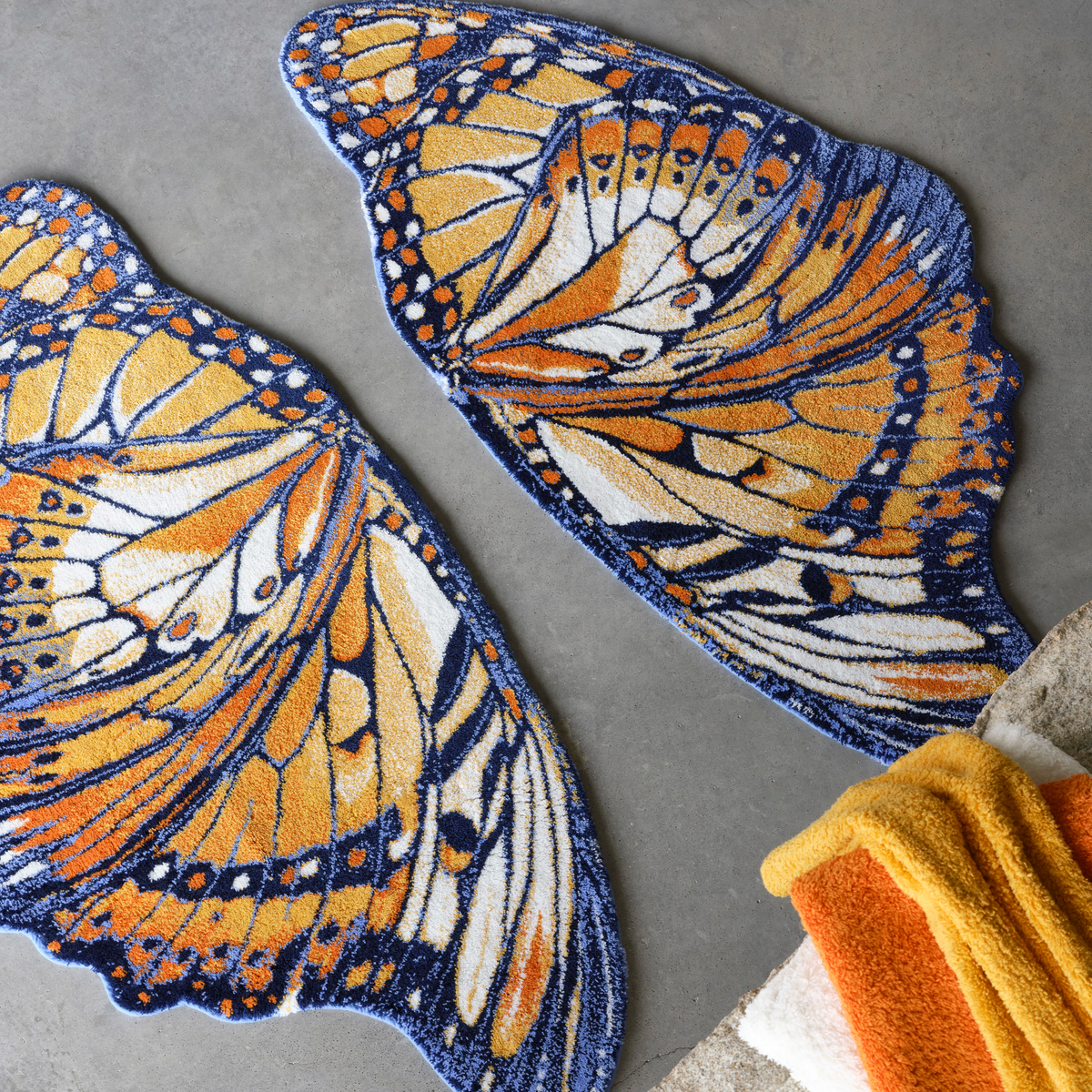 Two luxury Abyss Habidecor Mariposa bath rugs shaped like butterfly wings in orange, black, and white lie on a gray floor next to matching colored towels