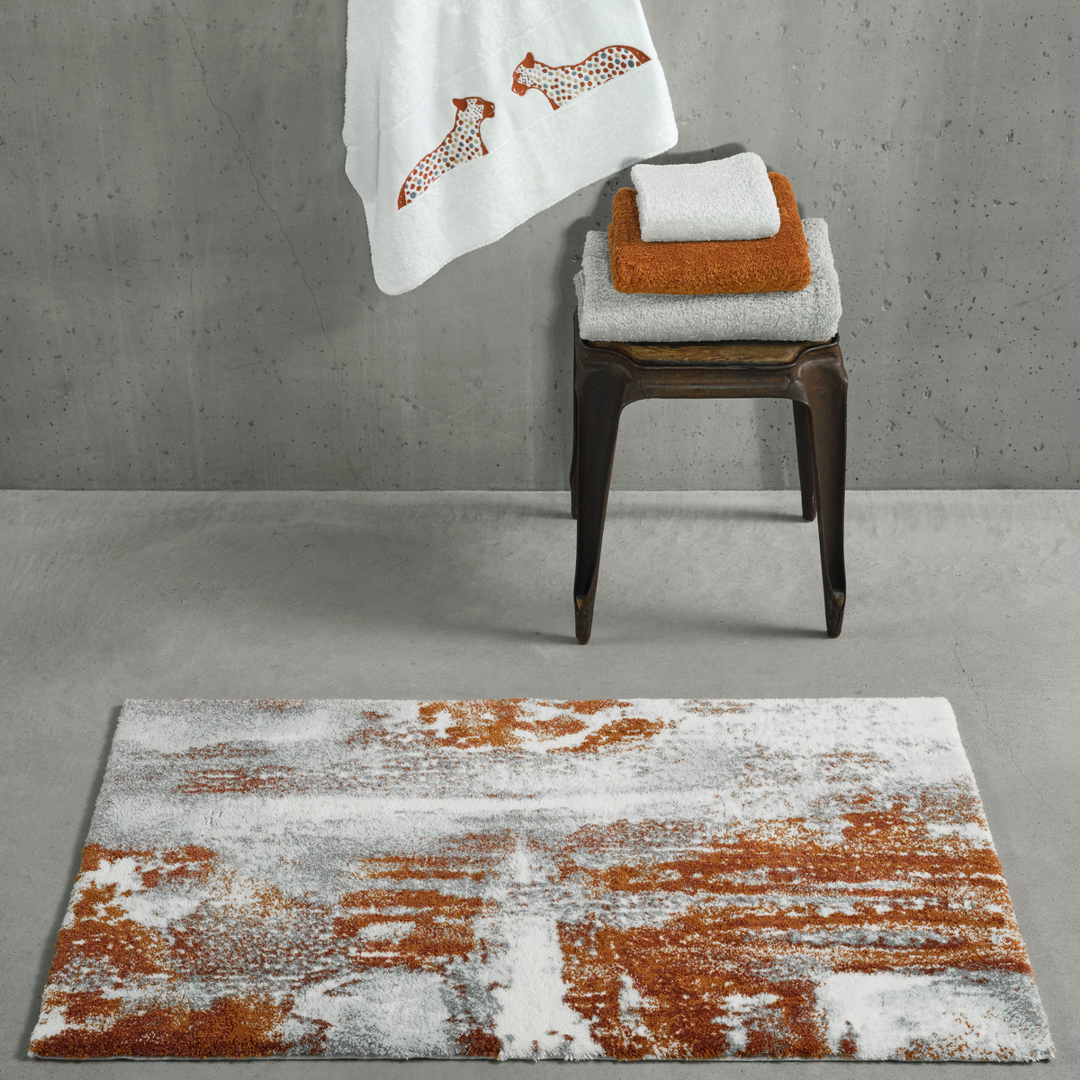 Caramel Abyss Habidecor Sparks Bath Rug on a concrete floor, with matching colored towels neatly arranged