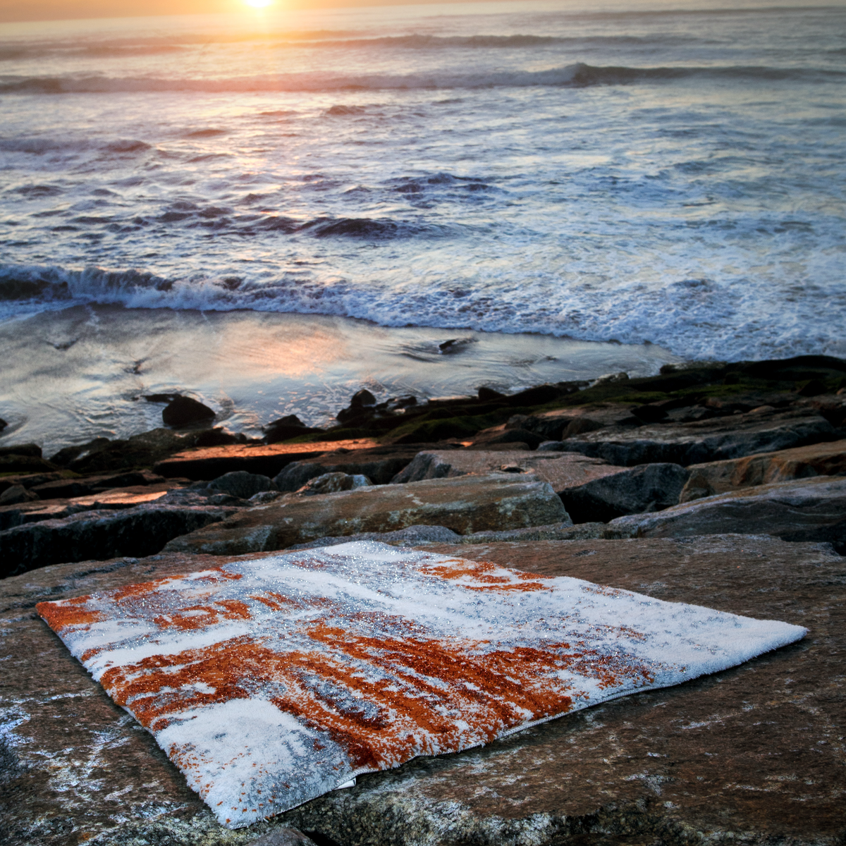 Caramel colored Abyss Habidecor Sparks Bath Rug lies on rocky ground by the ocean, waves and sunset in the background
