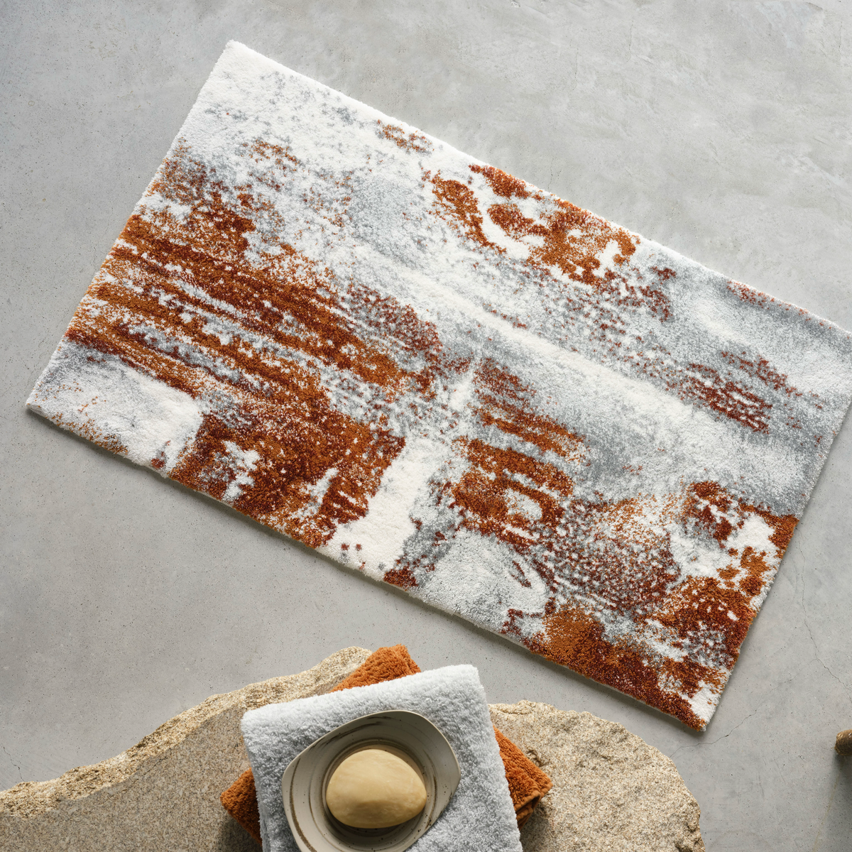 Caramel Abyss Habidecor Sparks Bath Rug lies on a light gray floor near towels and soap
