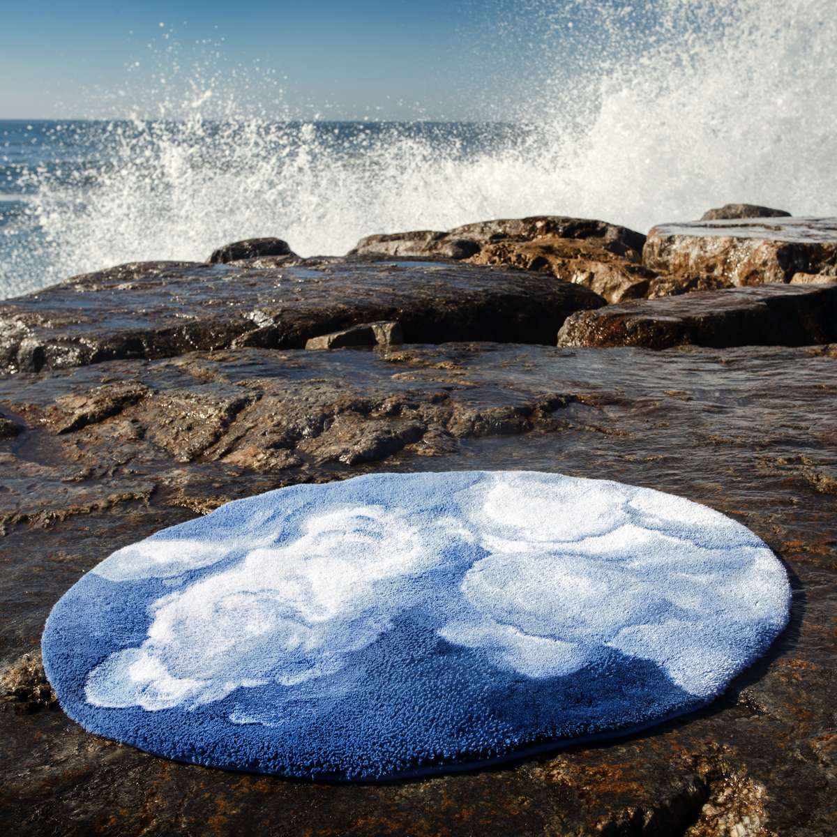 Regatta colored Abyss Habidecor Walker Bath Rug sits on a rocky shore as waves crash in the background