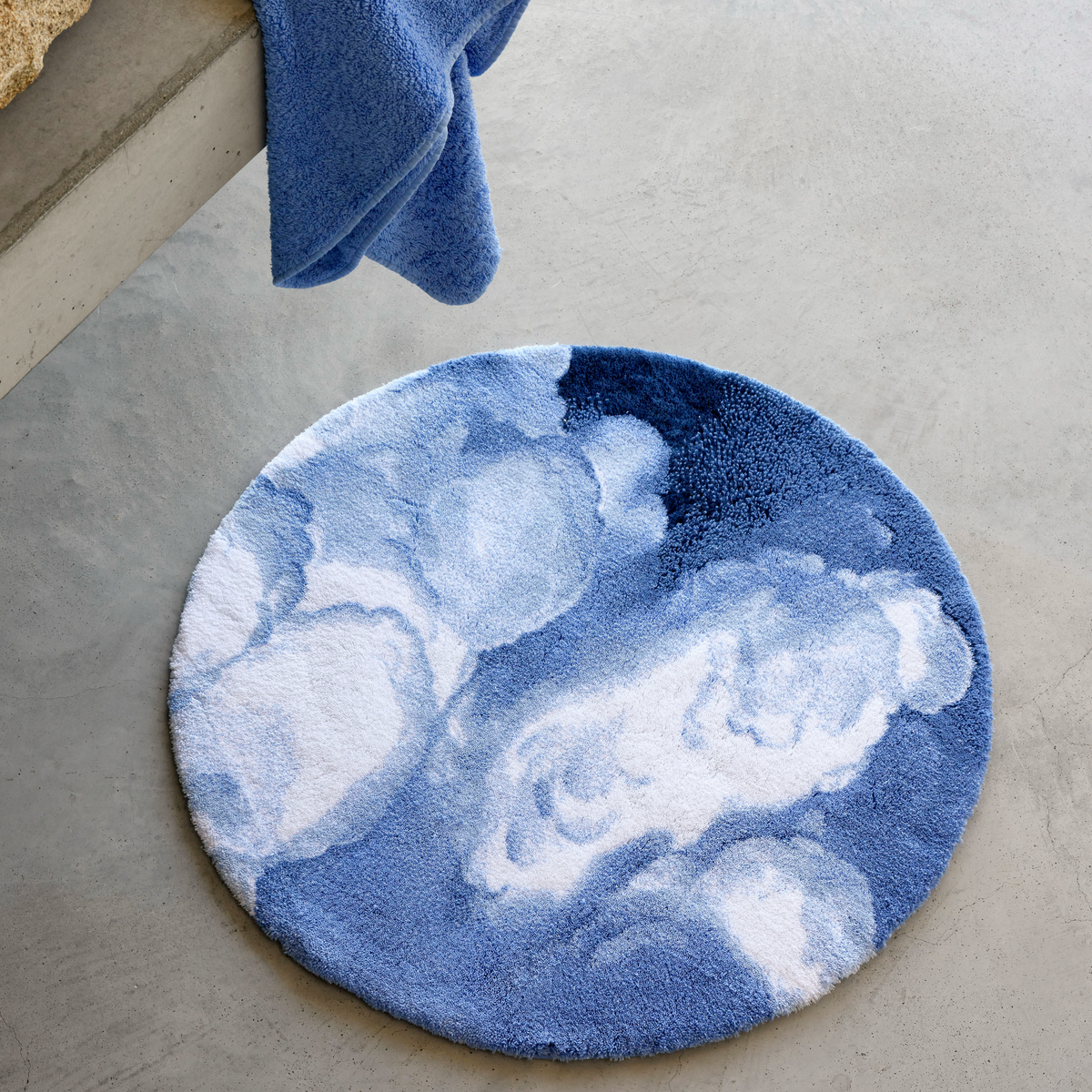 Abyss Habidecor Walker Bath Rug in blue and white clouds sits on a light gray concrete floor