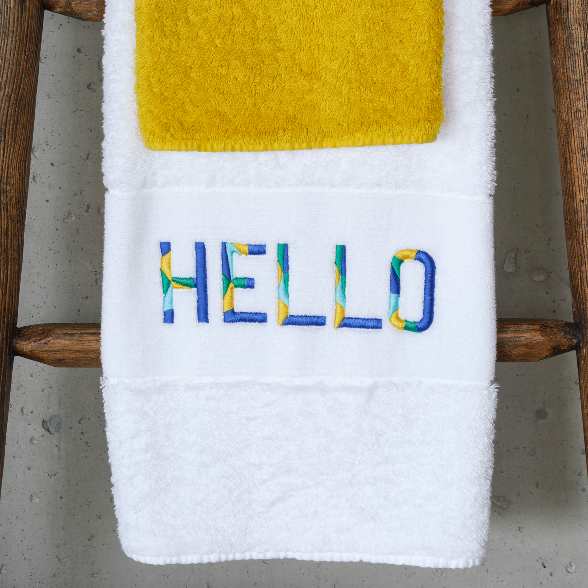 Abyss Hello Embroidered Bath Towels in Marina (304) color hangs below a yellow luxury towel on a wooden rack
