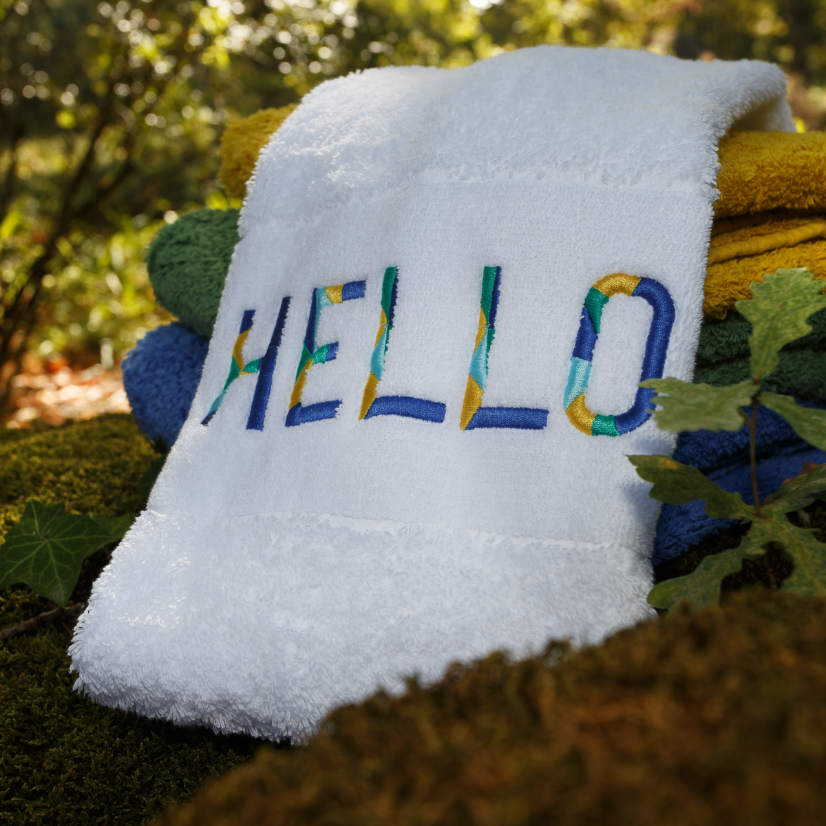 A luxury embroidered towel with HELLO in colorful letters tops a stack of towels outdoors.