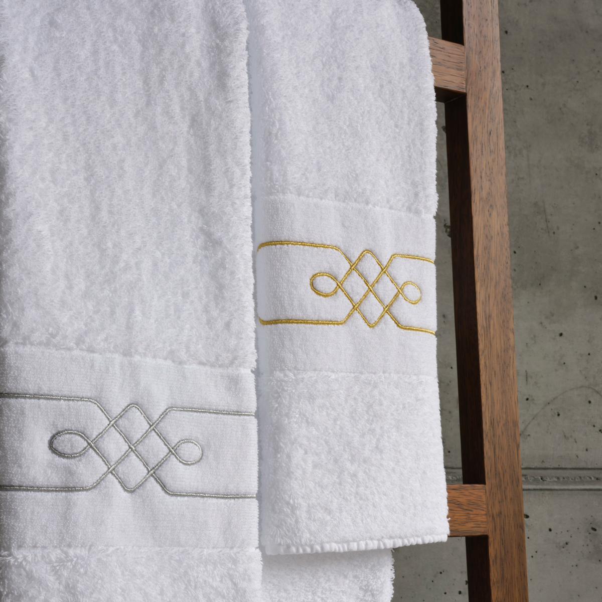 Two Abyss Lasso Towels with gold and silver embroidery hang on a wooden rack—luxury Egyptian Giza cotton towels.