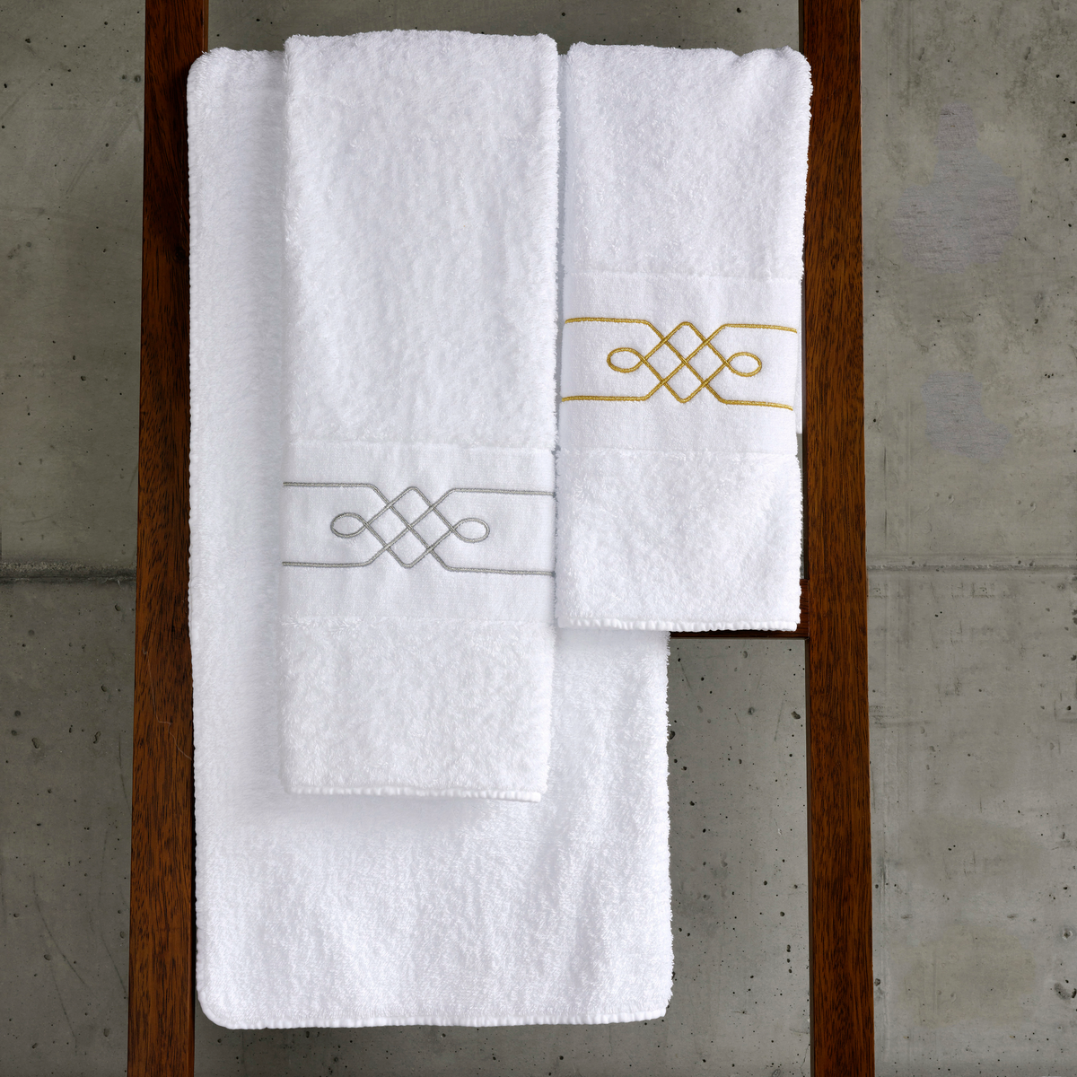 Three white luxury embroidered towels, one gray and one gold, are neatly stacked on a wooden rack.