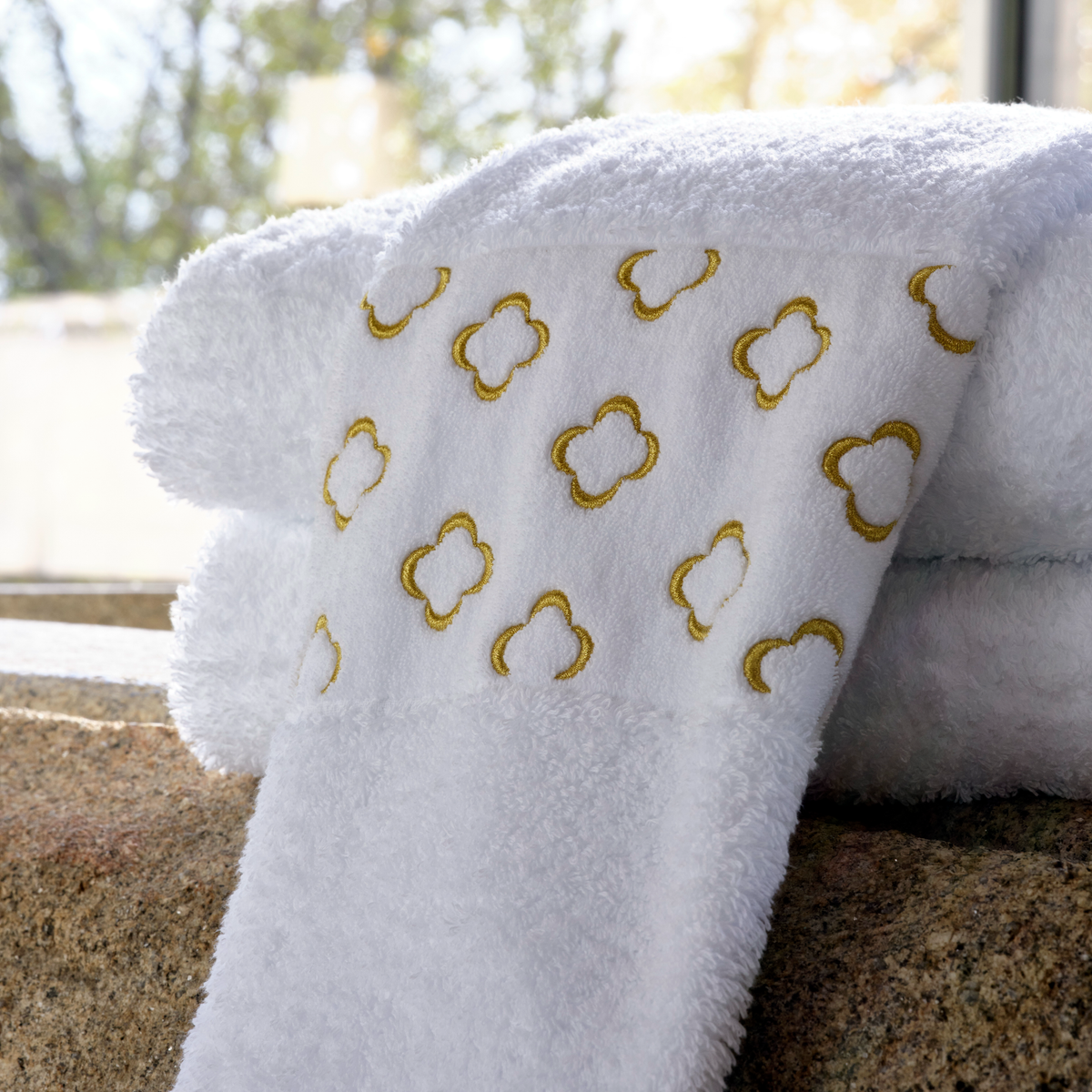 A white Abyss Luna Towel with a gold quatrefoil pattern is folded on a stone surface near a window.