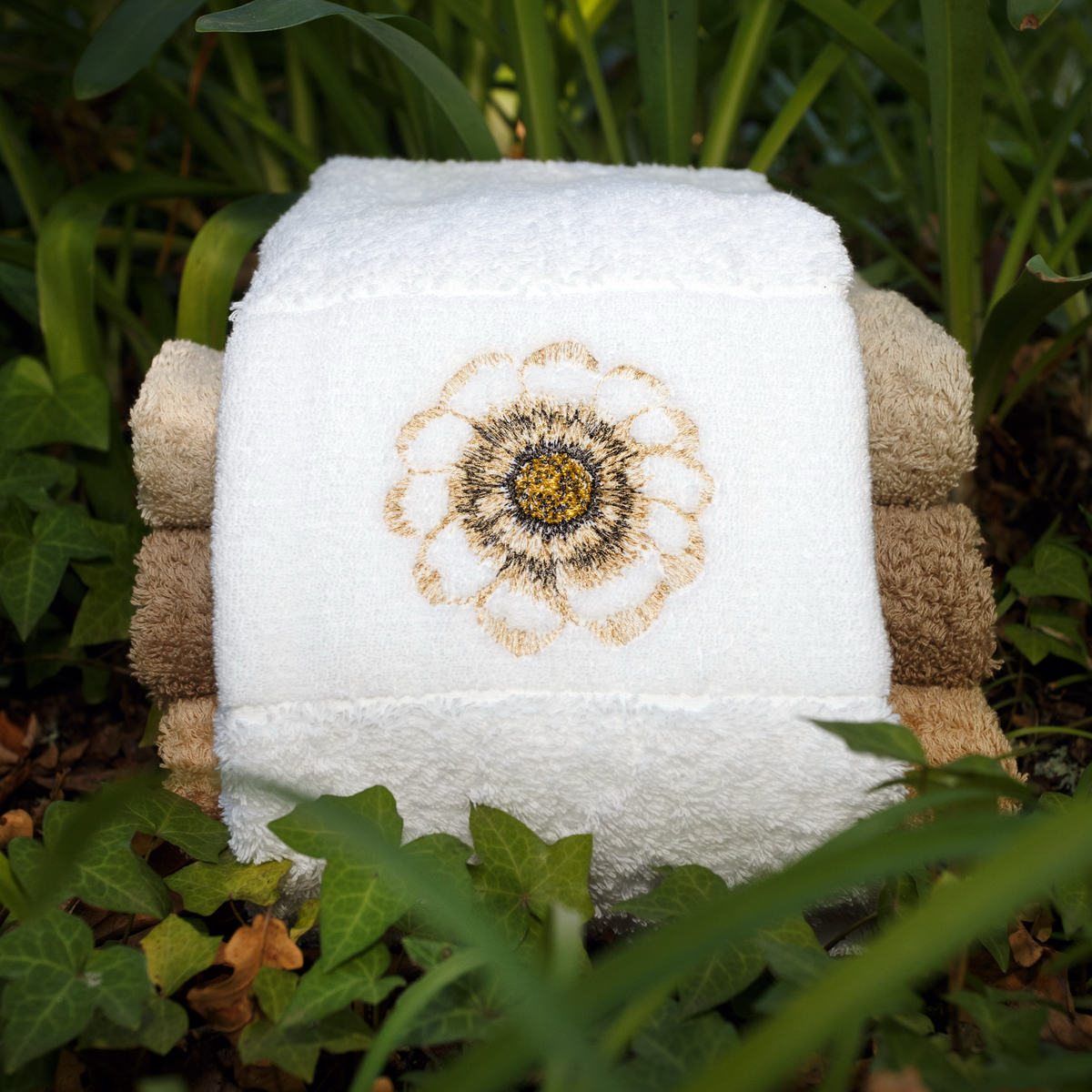 Three folded botanical embroidered towels, including an Egyptian cotton towel, stacked outdoors among green leaves.