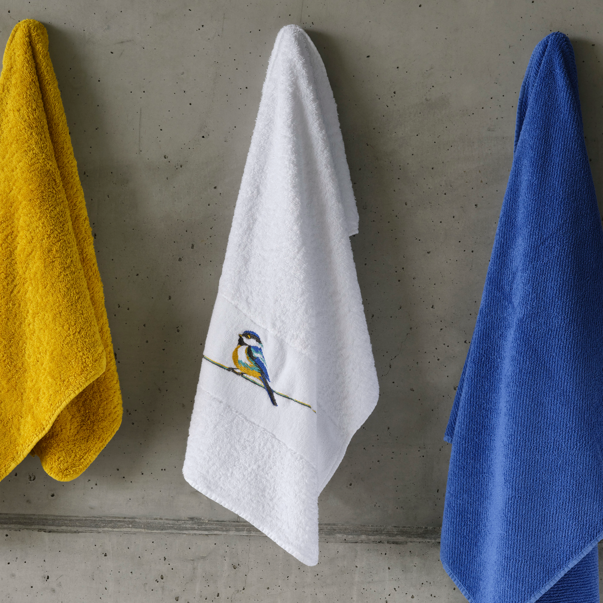 Three towels hang on a concrete wall: an Abyss Mesange Towel, an embroidered bird towel, and Egyptian Giza cotton blue.
