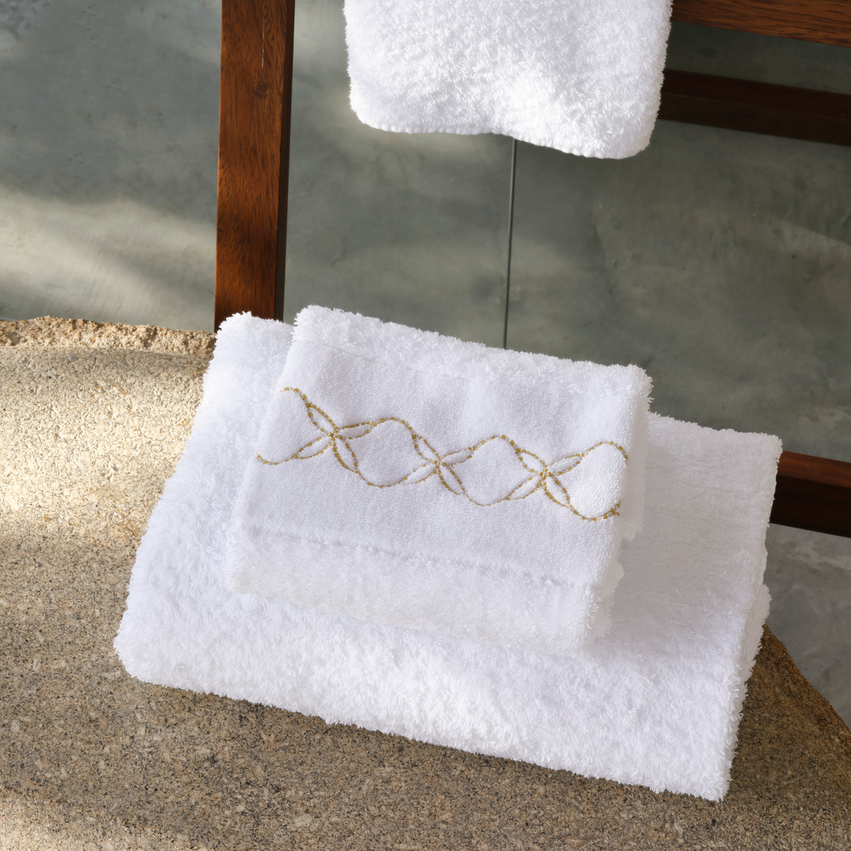 Two folded white luxury embroidered towels sit on a stone surface, one featuring a gold pattern.