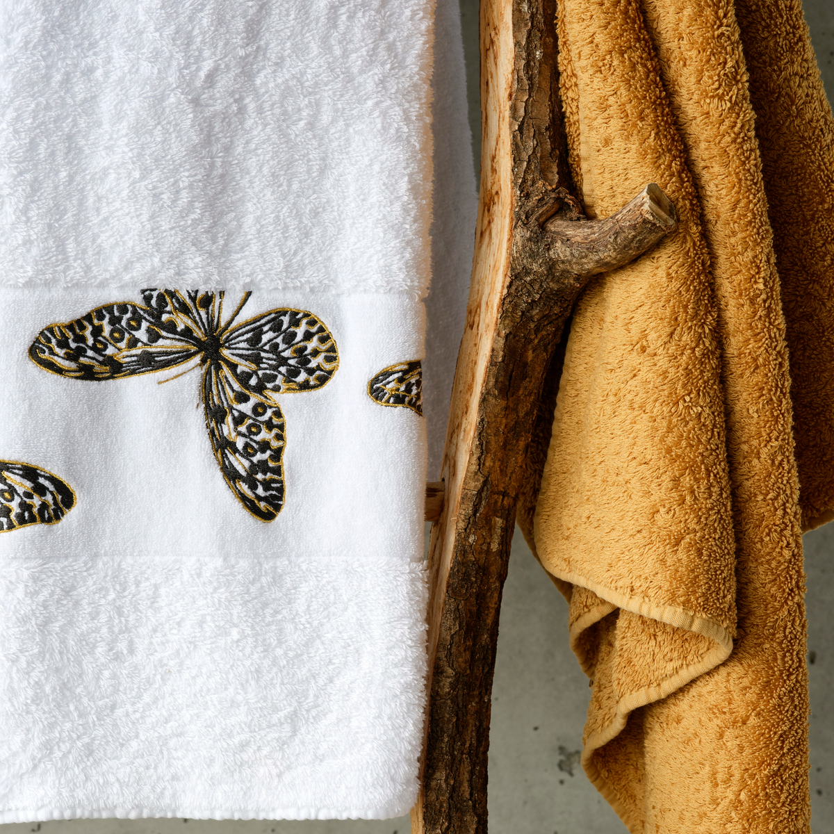 A butterfly embroidered towel and a luxury bath towel in mustard yellow hang side by side on a wooden hook.