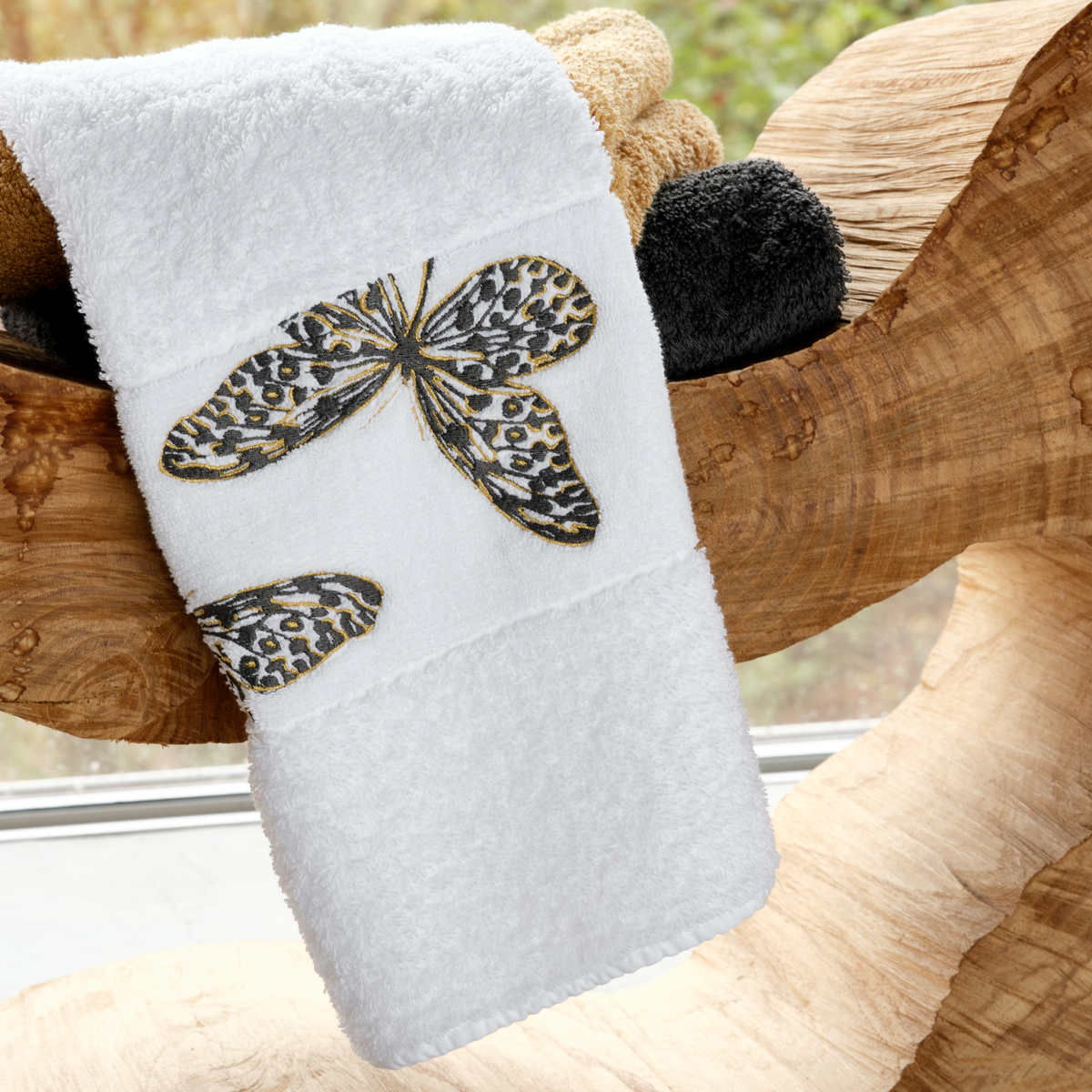 A butterfly embroidered towel in soft Egyptian cotton is draped over wood, with other luxury bath towels behind.