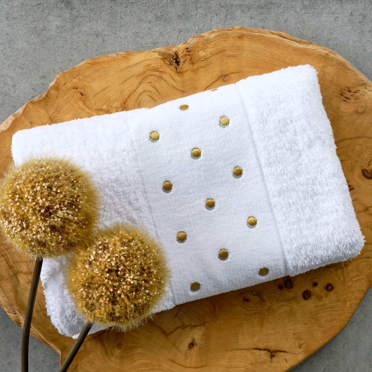 A metallic embroidered towel with gold polka dots rests on wood beside two dried flowers, crafted from Egyptian Giza cotton.