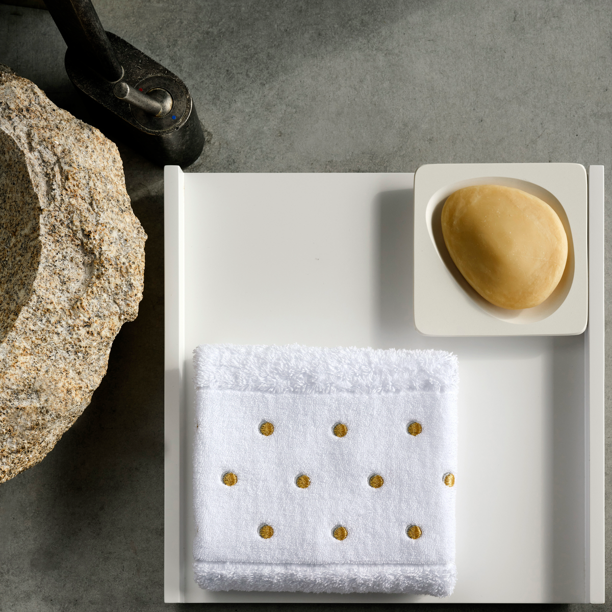 A white tray holds a bar of soap and an Abyss Pintas Towel, a metallic embroidered towel, beside a stone basin.