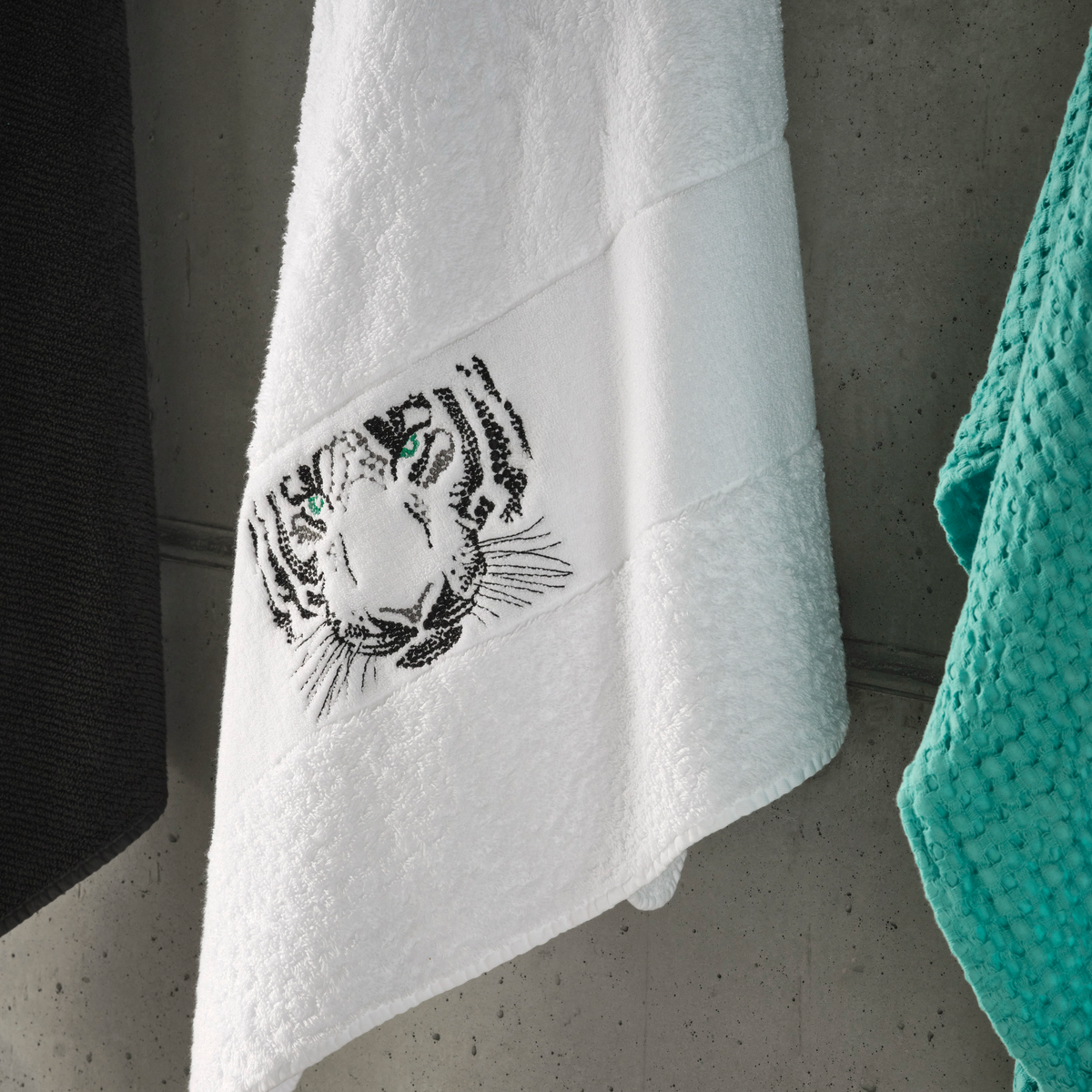 A white Abyss Tiger Towel in Egyptian Giza Cotton hangs beside a teal textured towel and a dark luxury towel.
