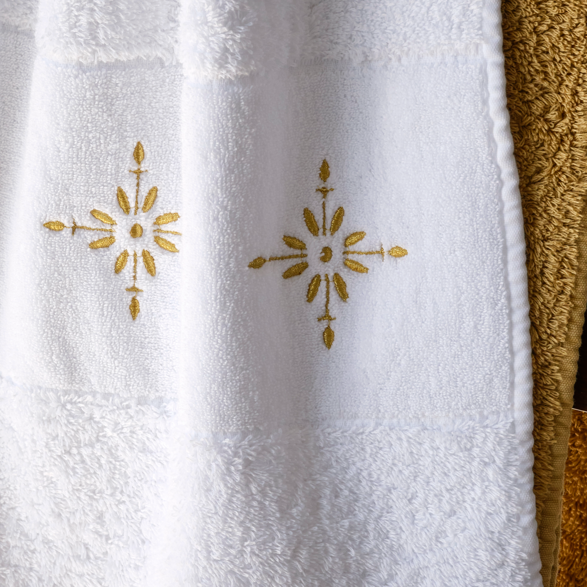A white Abyss Vega Towel with gold embroidery, crafted from Egyptian Giza Cotton, hangs by a textured tan towel.
