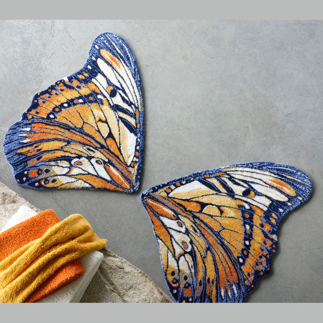 Two butterfly wing-shaped luxury bath rugs in blue, yellow, and orange lie on a gray floor near folded towels.