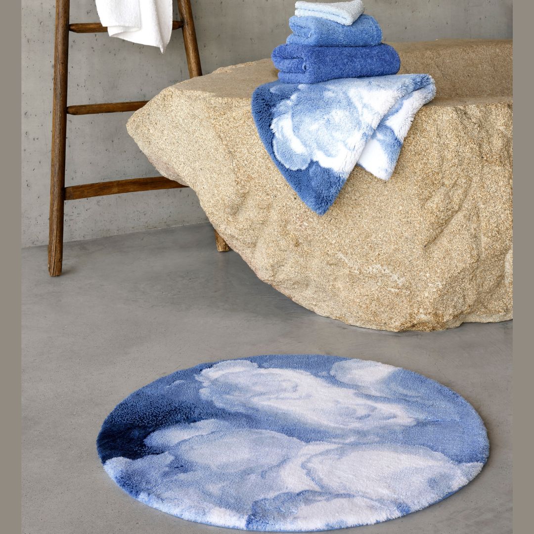 A plush absorbent bath rug in blue and white lies on a gray floor near towels and blankets on a tan stone bench.