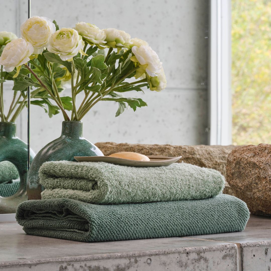 Two Abyss Twill Bath Towels stacked on a bathroom counter with a vase of white flowers and a bar of soap on a dish.