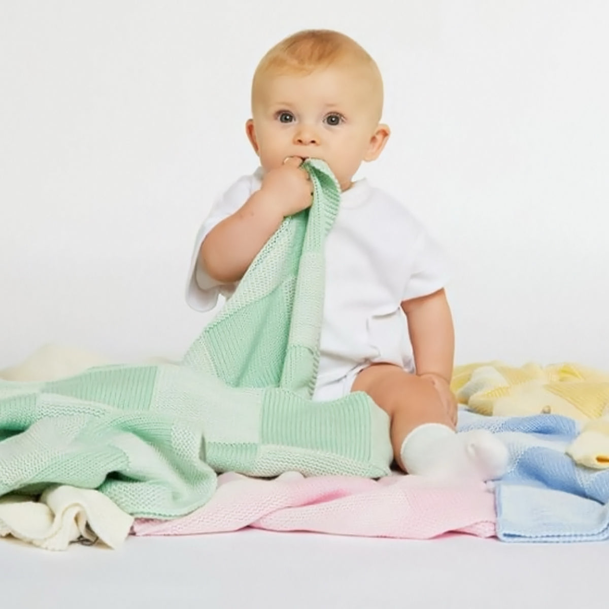 A baby in a white onesie sits on pastel blankets, chewing on a soft cashmere baby blanket.