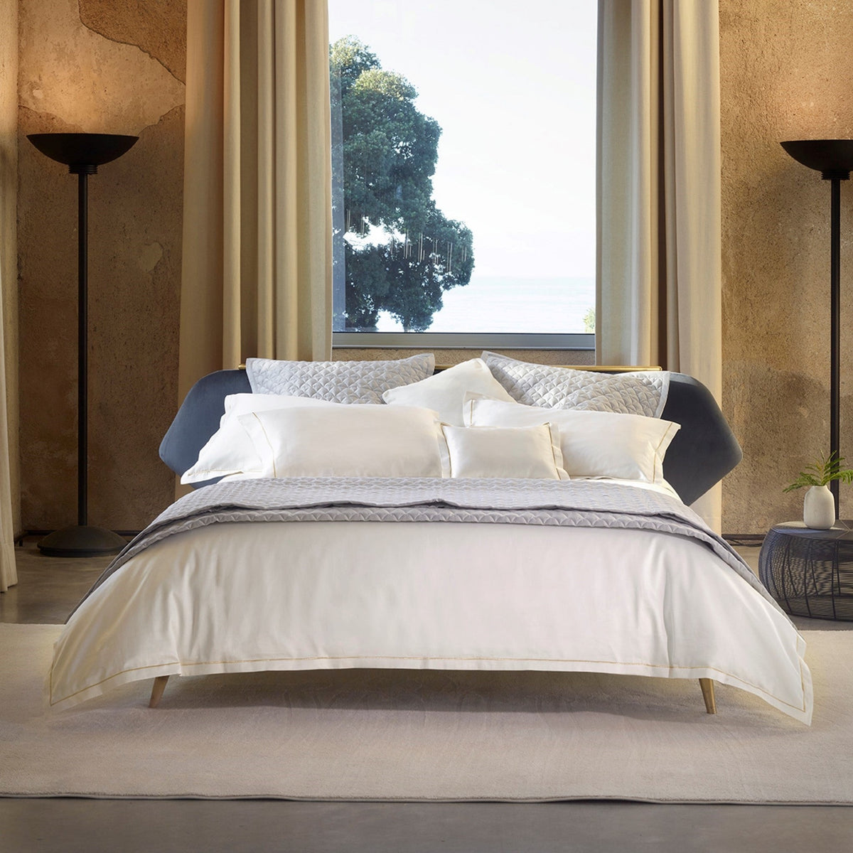A bed with layered white and grey Giza 92 Cream Gold Amalia Dalia Bedding centered in front of a window.