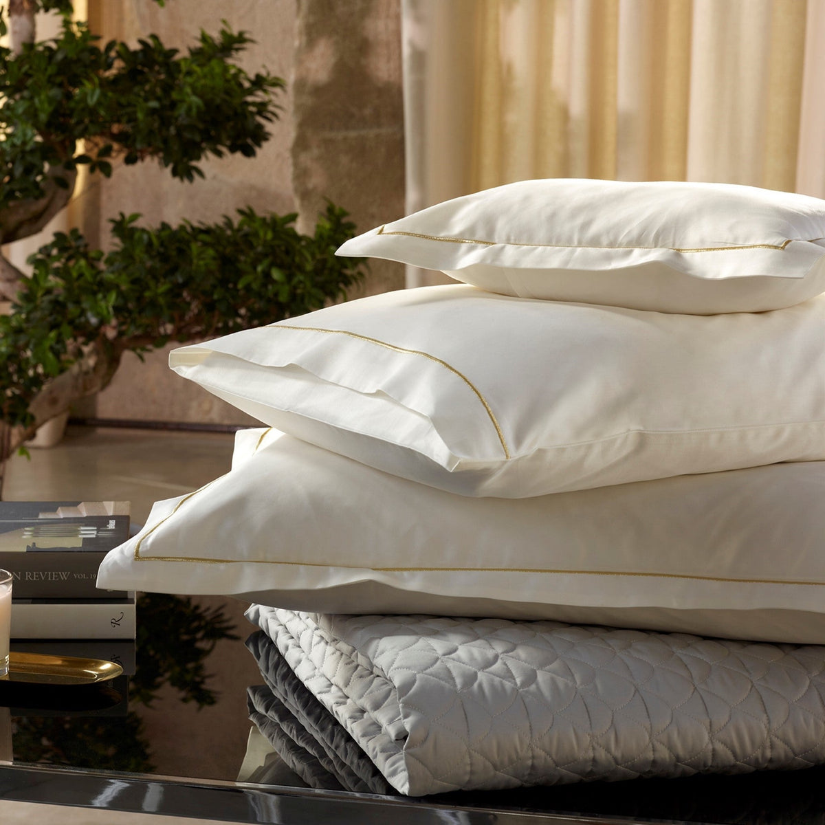 A stack of three Egyptian cotton sateen Cream Gold Amalia Dalia Shams and Pillow resting on a folded quilted grey blanket.