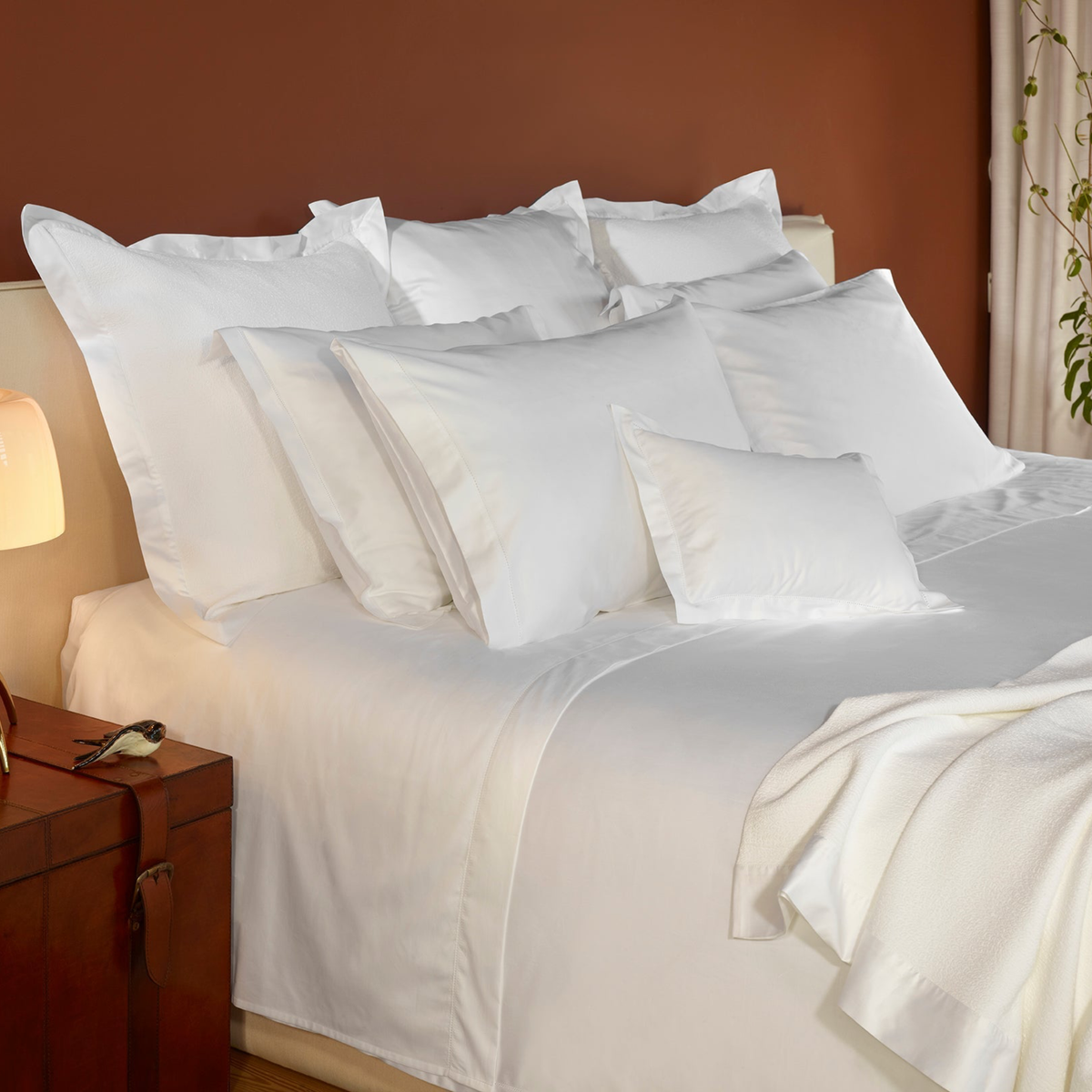 A bed made with 430 thread count White Amalia Suave Giza 94 Bedding, next to a brown leather nightstand with a lamp