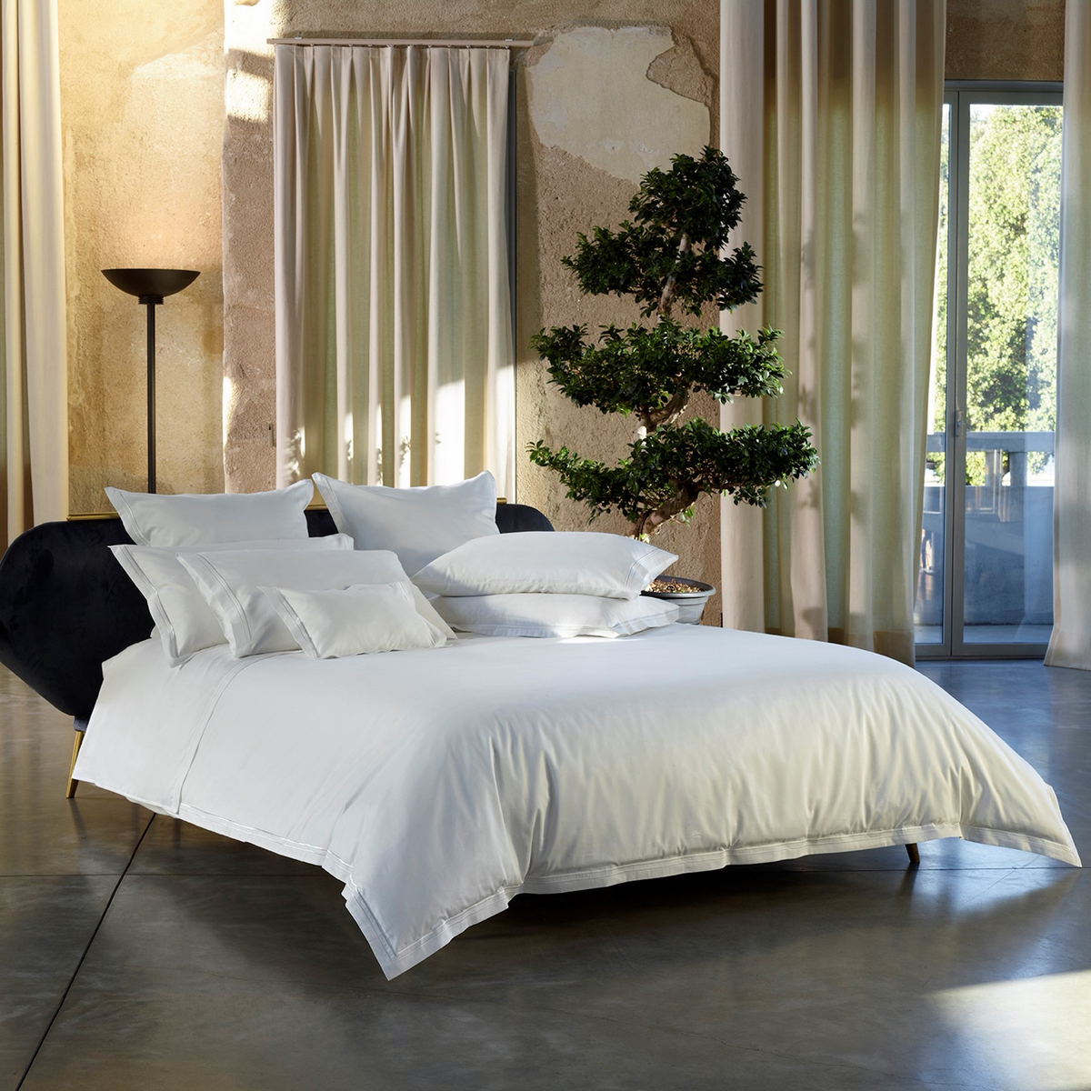 A neatly made bed with White Amalia Sublime Giza 45 Cotton Bedding, set by a decorative tree and floor-to-ceiling curtains