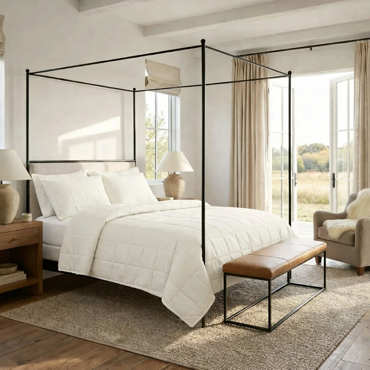 A bright bedroom with a four-poster bed, Bone colored Ann Gish Cotton Canvas Coverlet Set, a bench, two lamps, an armchair, and large windows