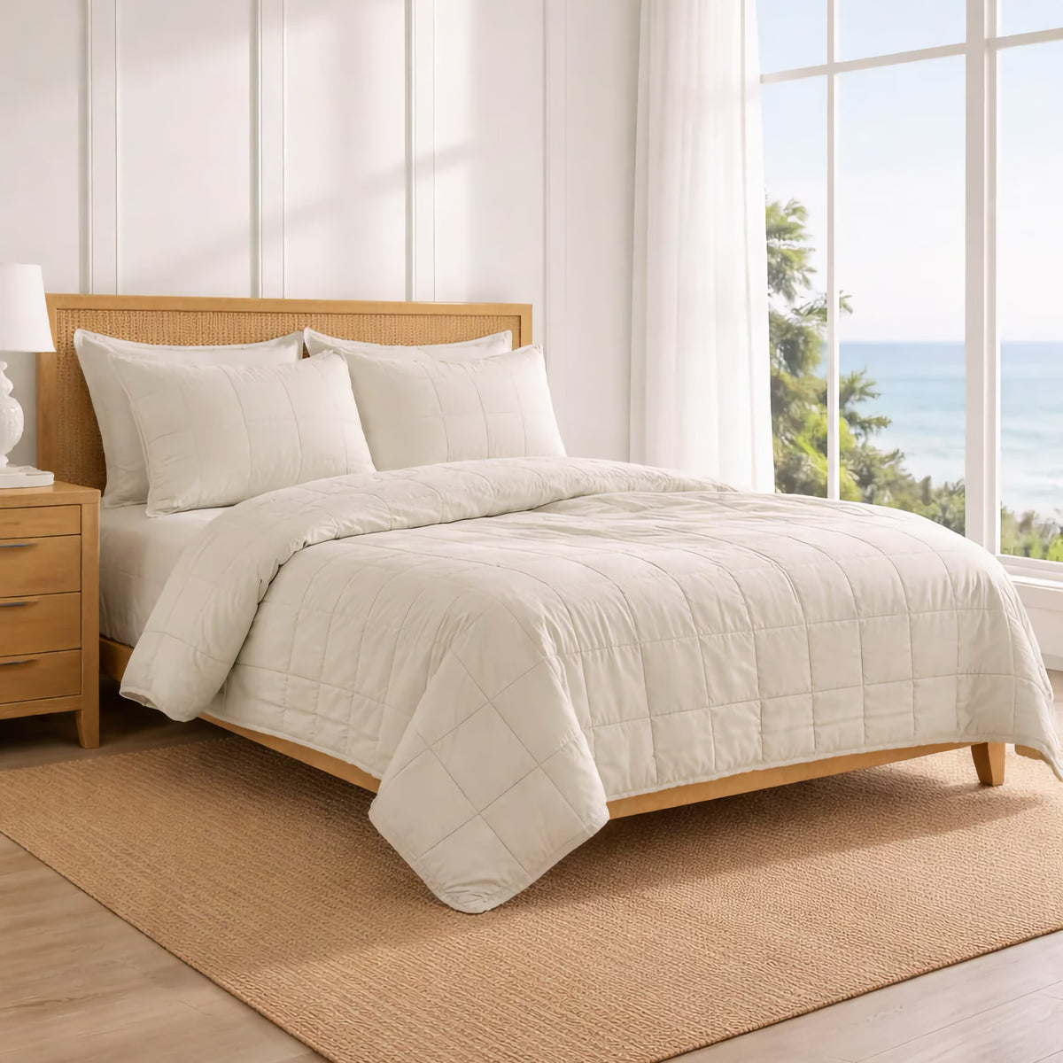 A neatly made bed with Bone colored Ann Gish Cotton Canvas Coverlet Set in a bright room and an ocean view through a large window