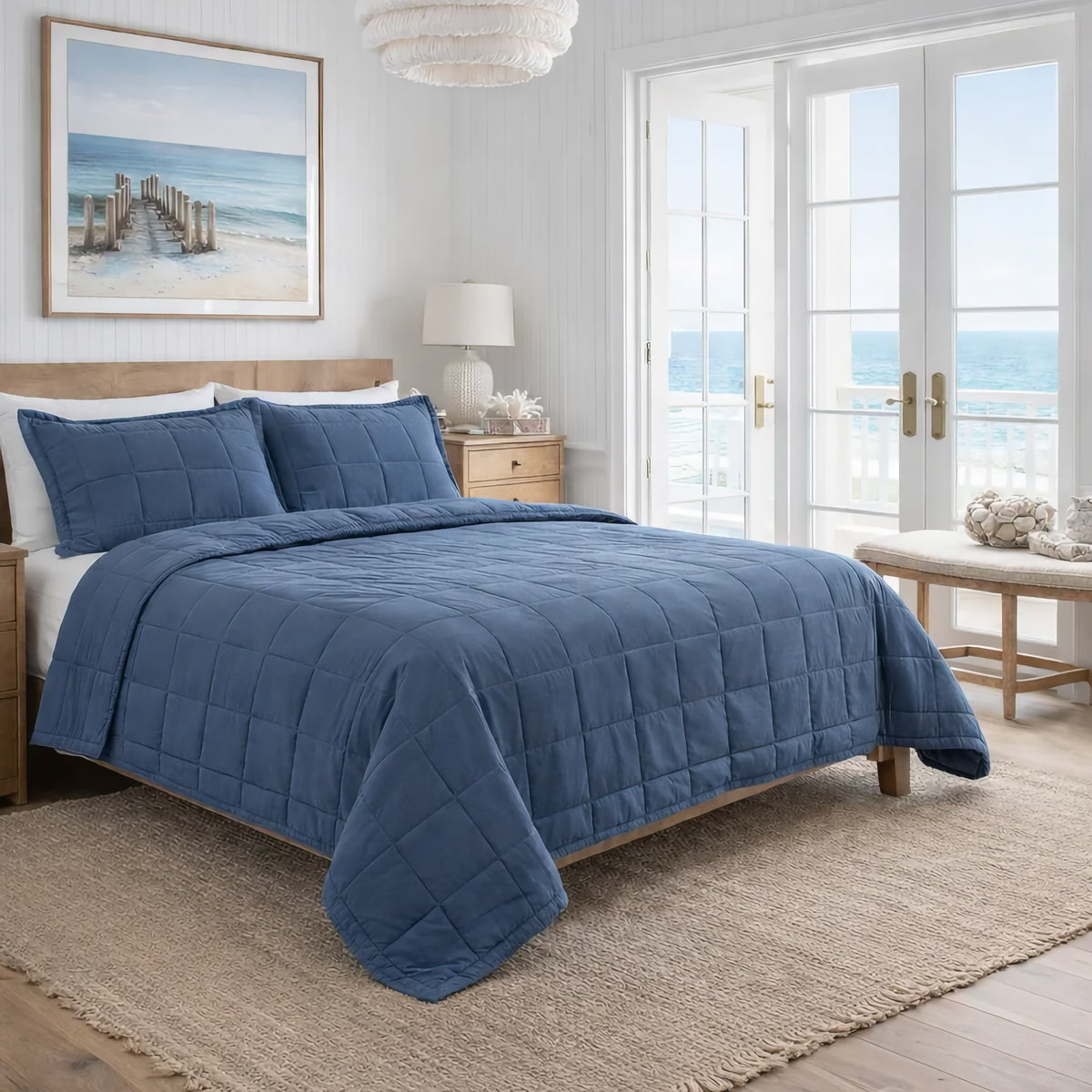 A neatly made bed with Denim colored Ann Gish Cotton Canvas Coverlet Set in a bright room and an ocean view through a large window