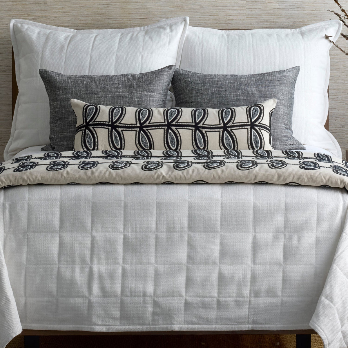 A neatly made bed with white bedding, and a Ann Gish Fellini Decorative Pillow in Natural and Charcoal colors
