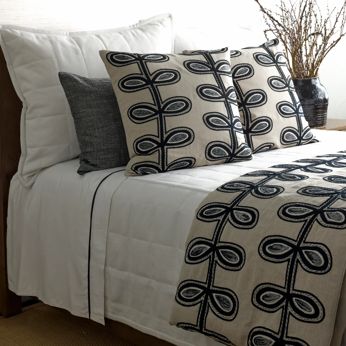 A bed with white bedding, and Ann Gish Fellini Throw and Decorative Pillows in Natural and Charcoal colors