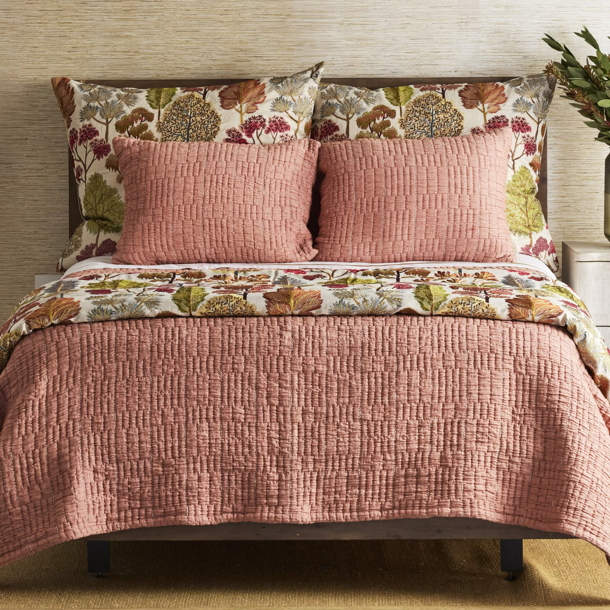 Bed with a textured Terra Cotta colored Ann Gish Pickstitch Coverlet Set, and floral shams and top sheet against a neutral wall