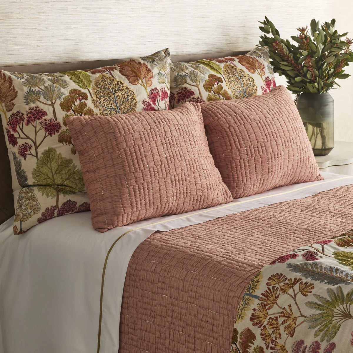 Closeup of Bed with a textured Terra Cotta colored Ann Gish Pickstitch Coverlet Set, and floral shams and top sheet against a neutral wall