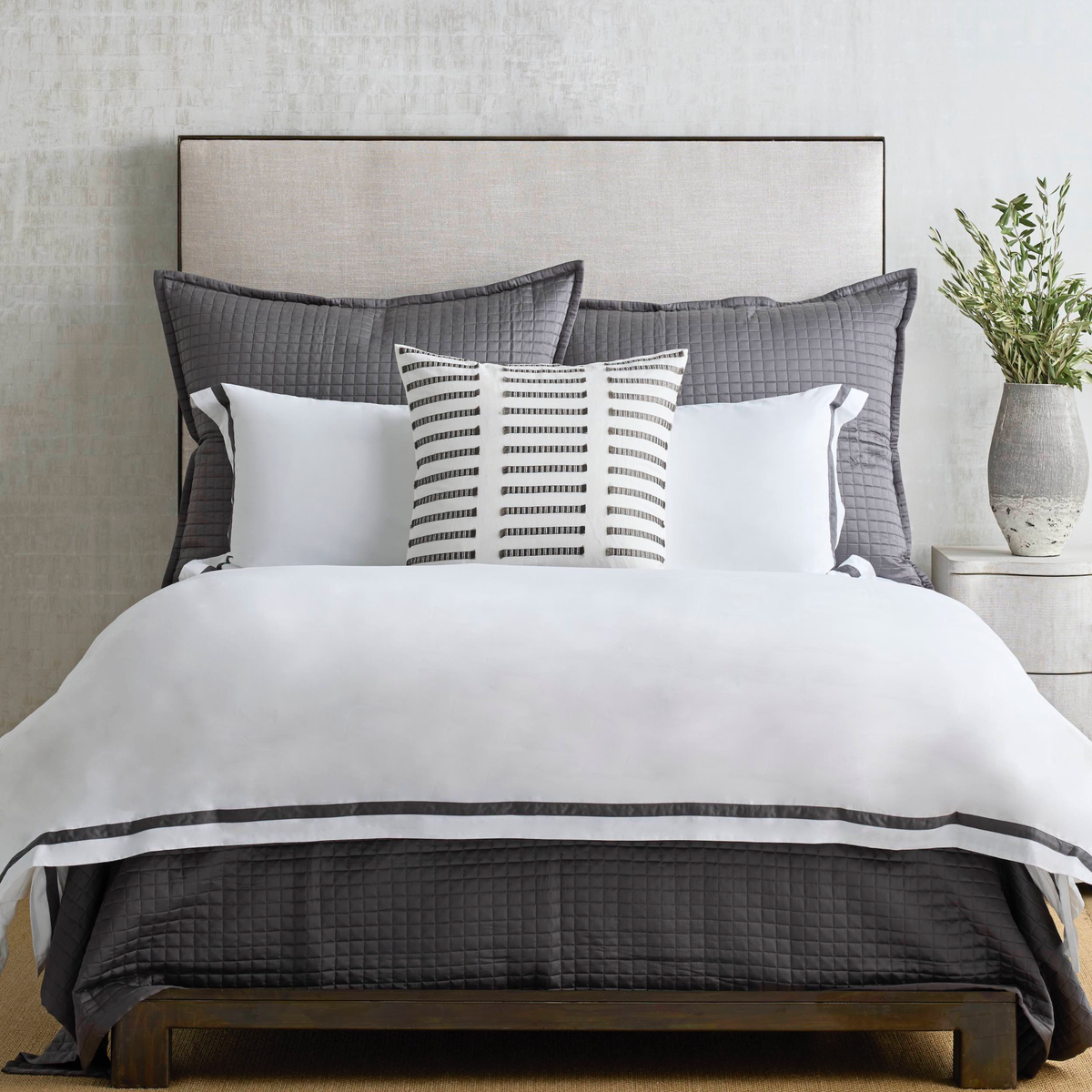 A made bed with gray quilted bedding, white pillows, a striped accent pillow, and a vase with greenery on a nightstand.