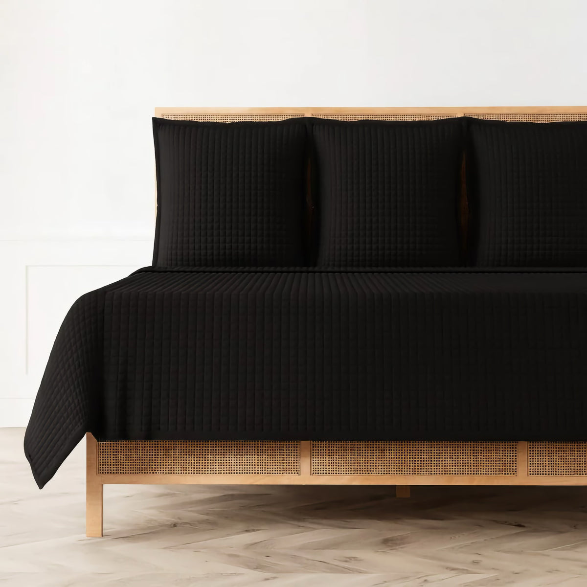 A bed with a wooden frame and an Ann Gish quilted coverlet, plus black pillows, creates a luxury bedding set against a white wall.
