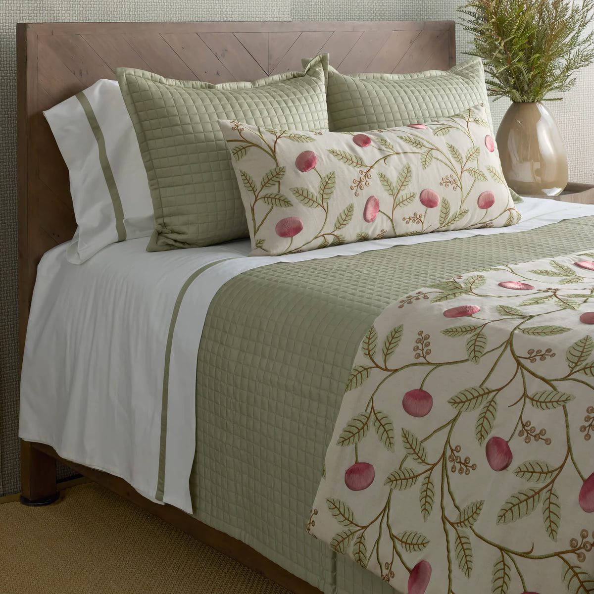 A neatly made bed with green Ann Gish quilted coverlet, white sheets, floral pillows, and a matching throw blanket.