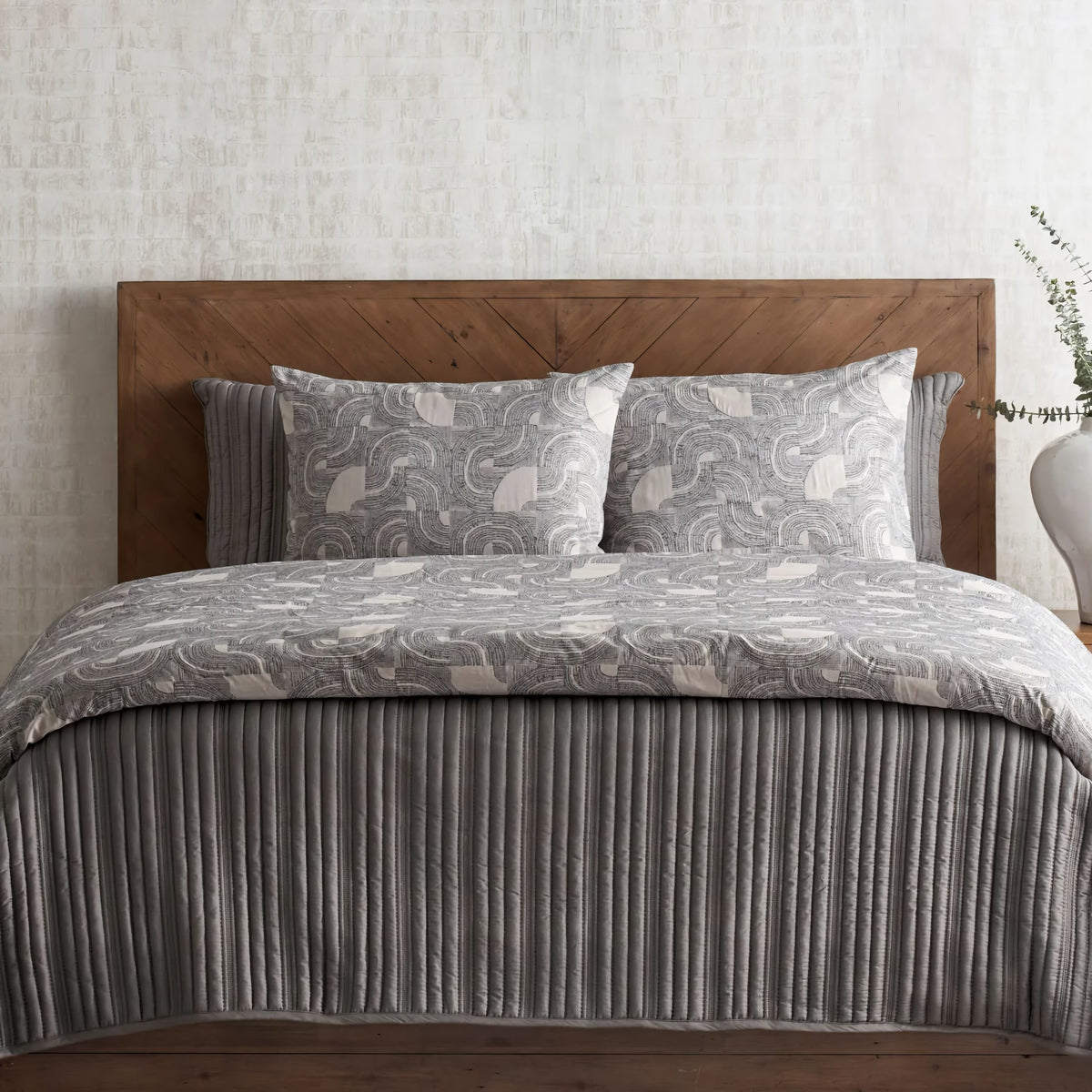 A neatly made bed with a wooden headboard, modern bedding, and a white vase with greenery on the right side.