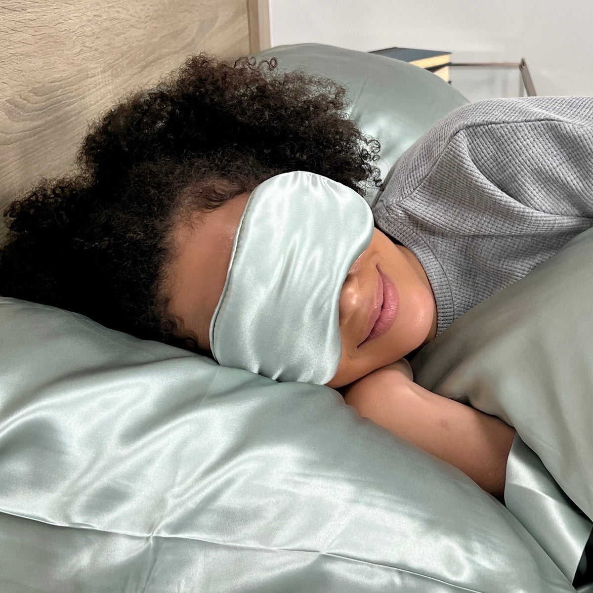 Woman relaxing on bed, wearing Sage silk sleep mask with matching silk pillows and sheet