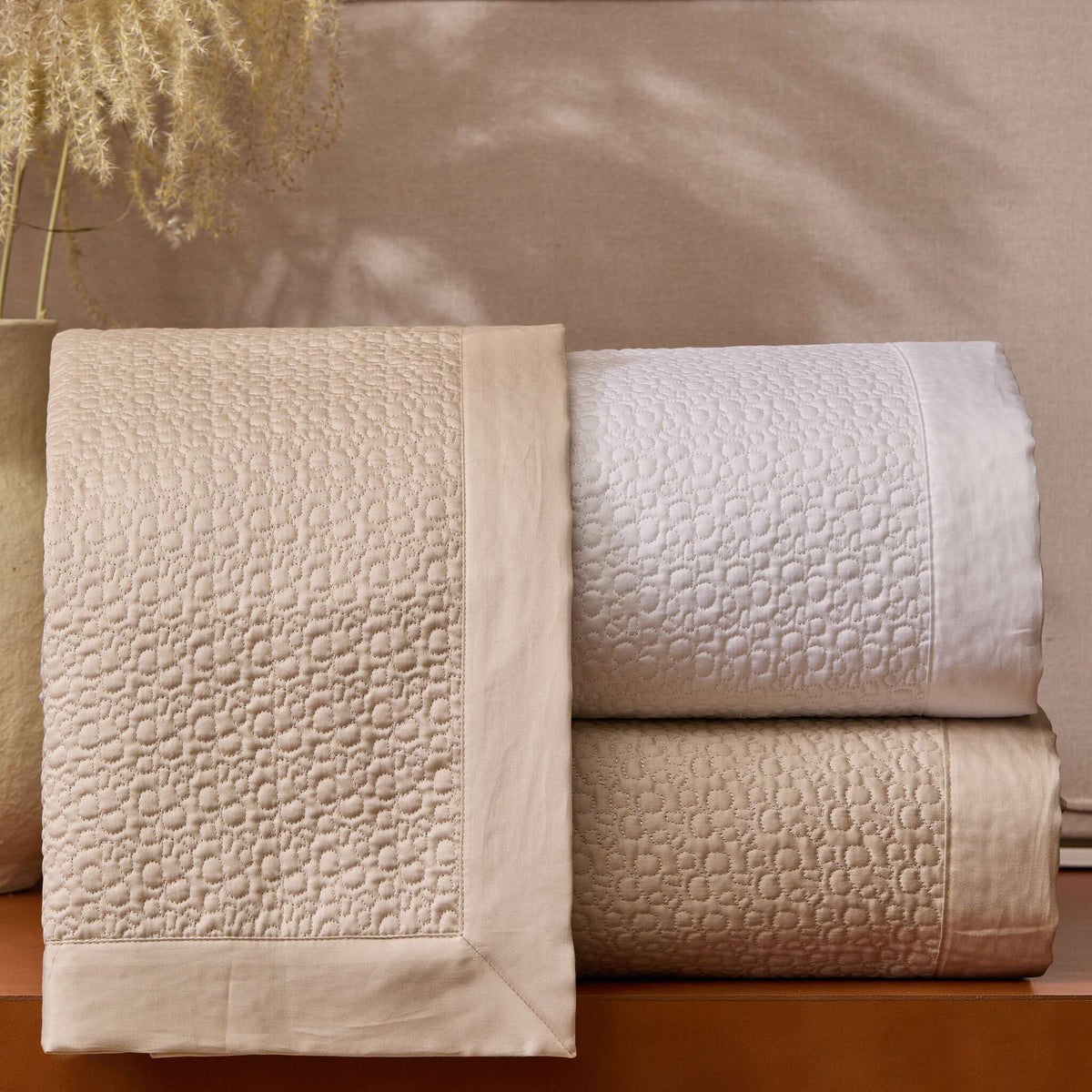 Bovi Estella Coverlet in White and Latte, stacked and displayed on a table with a plant in the background