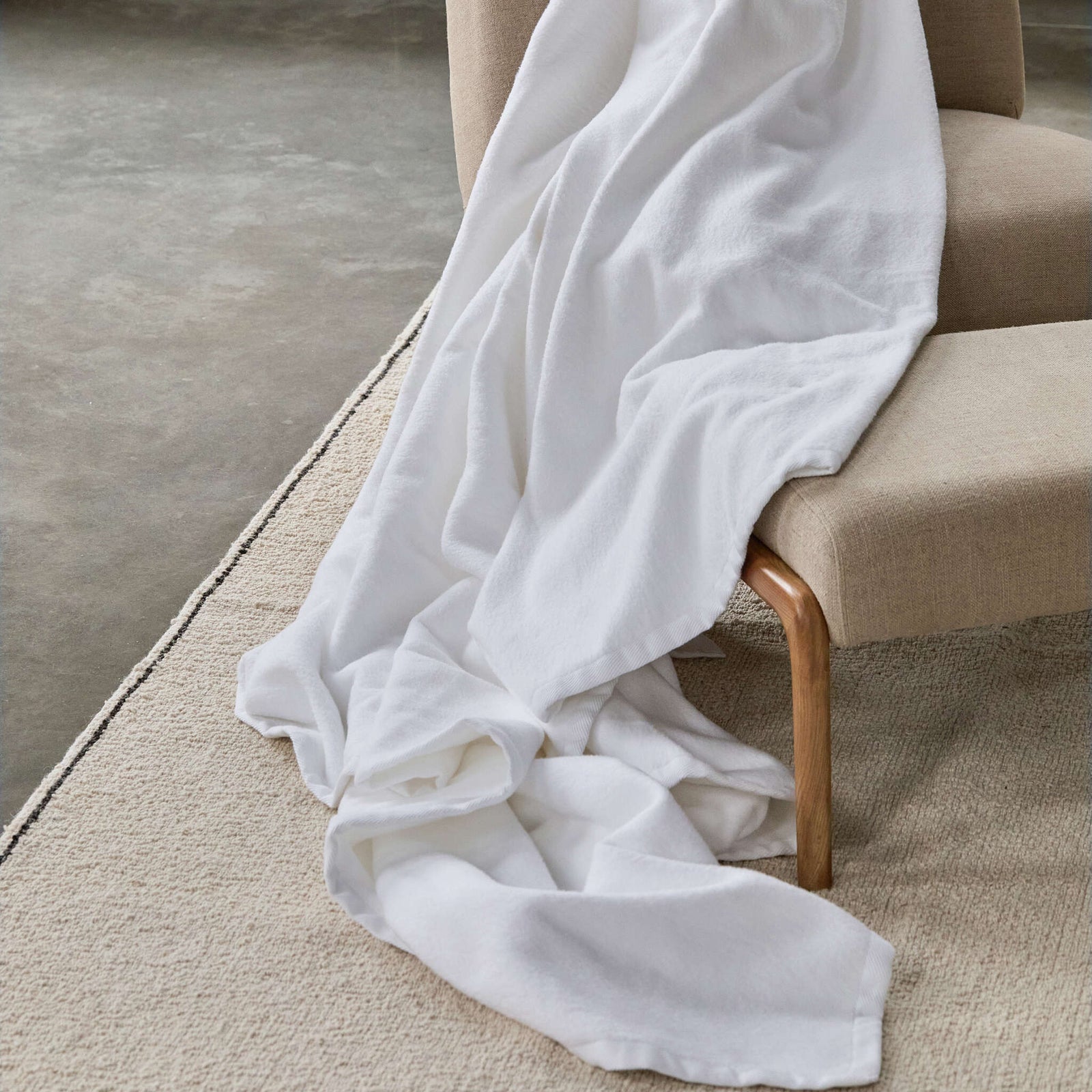White BOVI Finnley blanket draped over a beige chair on a carpeted floor
