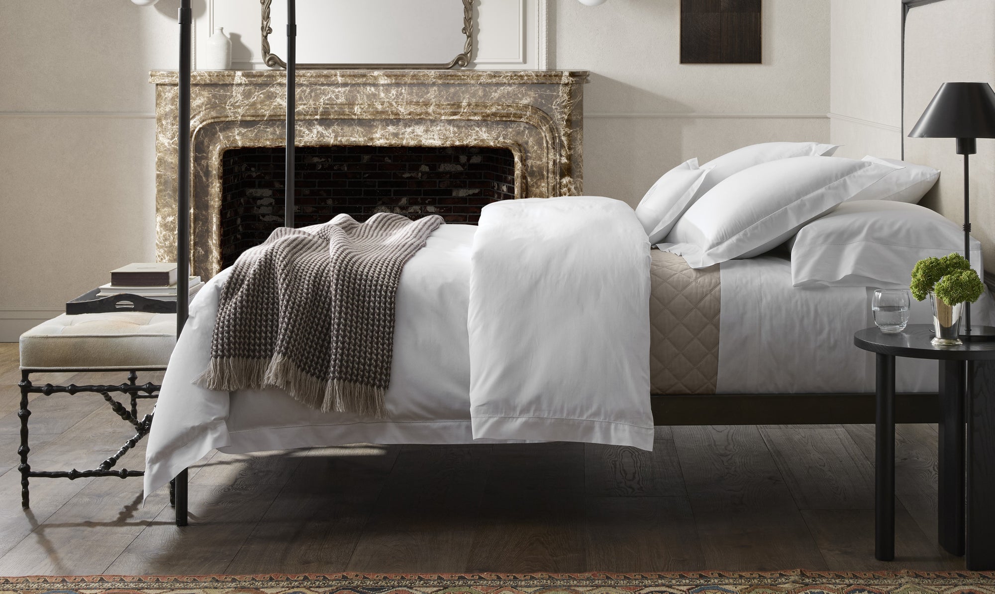 A neatly made bed with white linens and a brown throw blanket in a stylish bedroom with a marble fireplace.