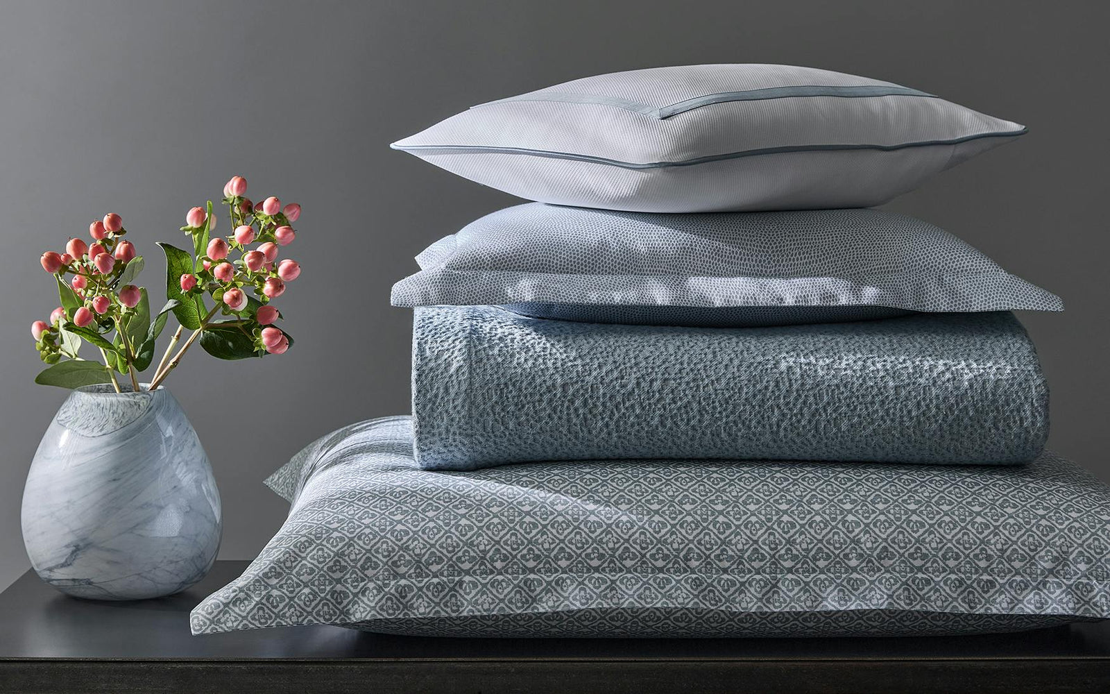 A marble vase with pink flowers sits next to a stack of blue and white patterned bedding and pillows against a gray wall.