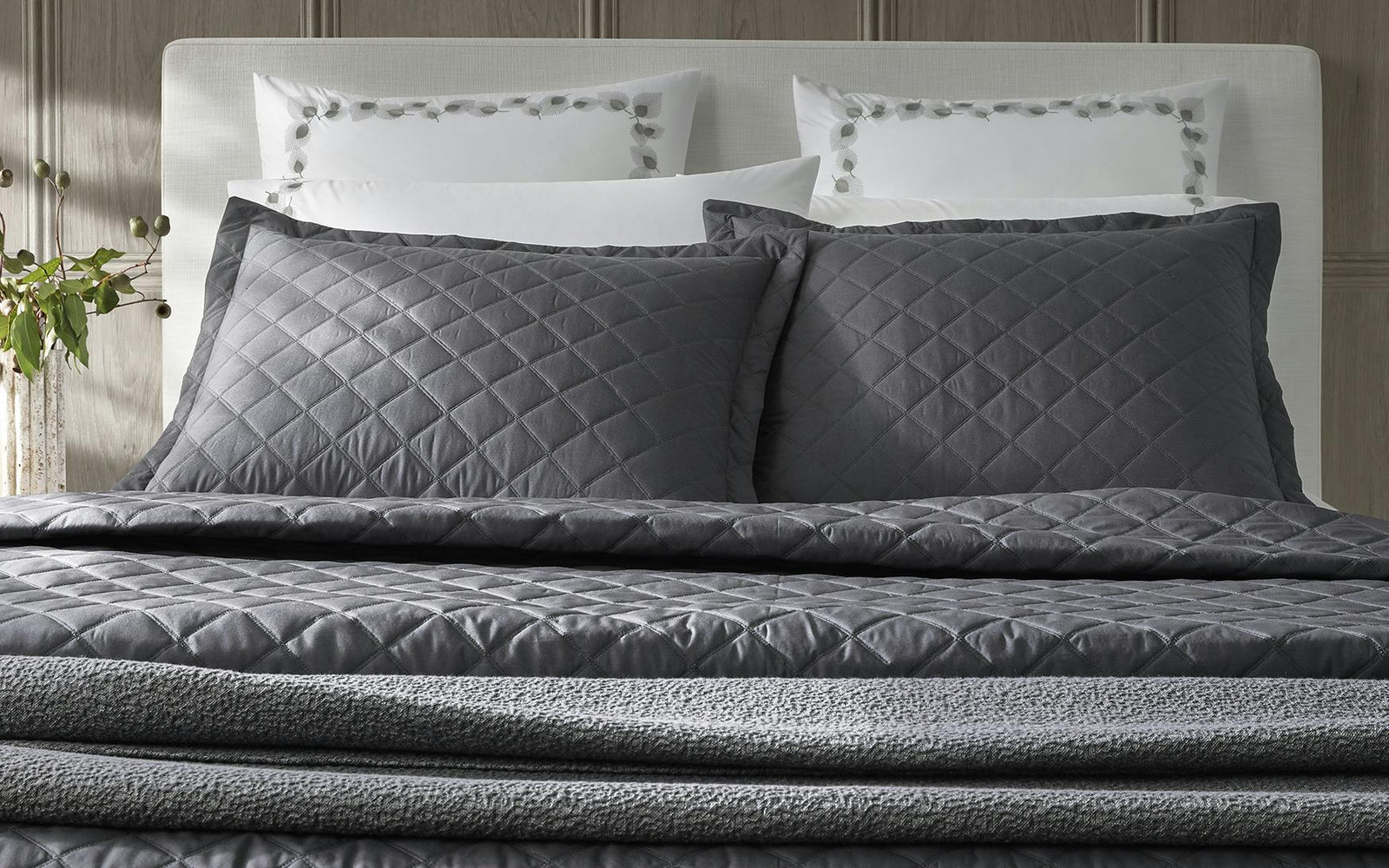 A neatly made bed with two gray quilted pillows and a matching comforter, white pillows in the background.