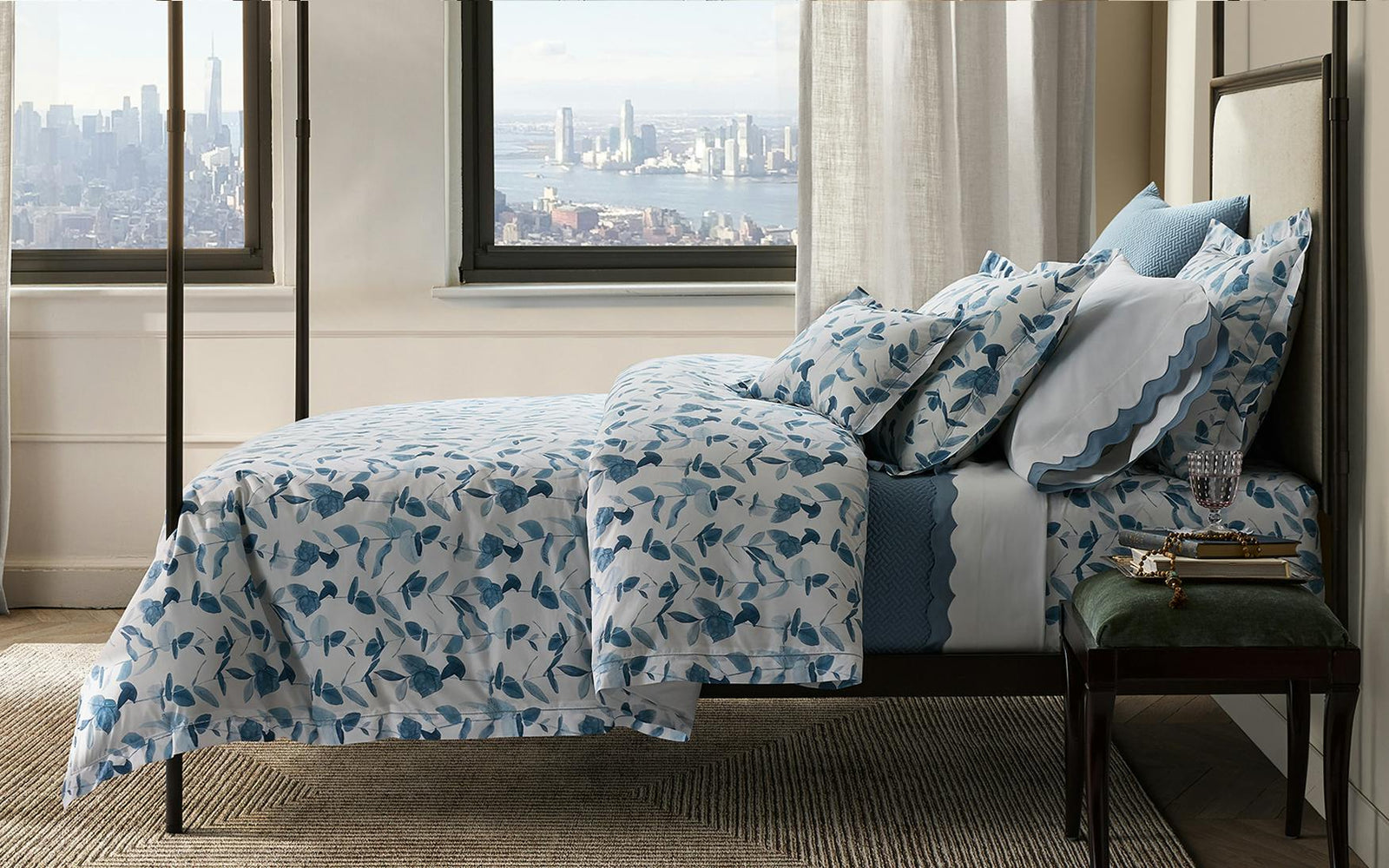 A neatly made bed with blue and white floral bedding is placed by a window overlooking a city skyline.